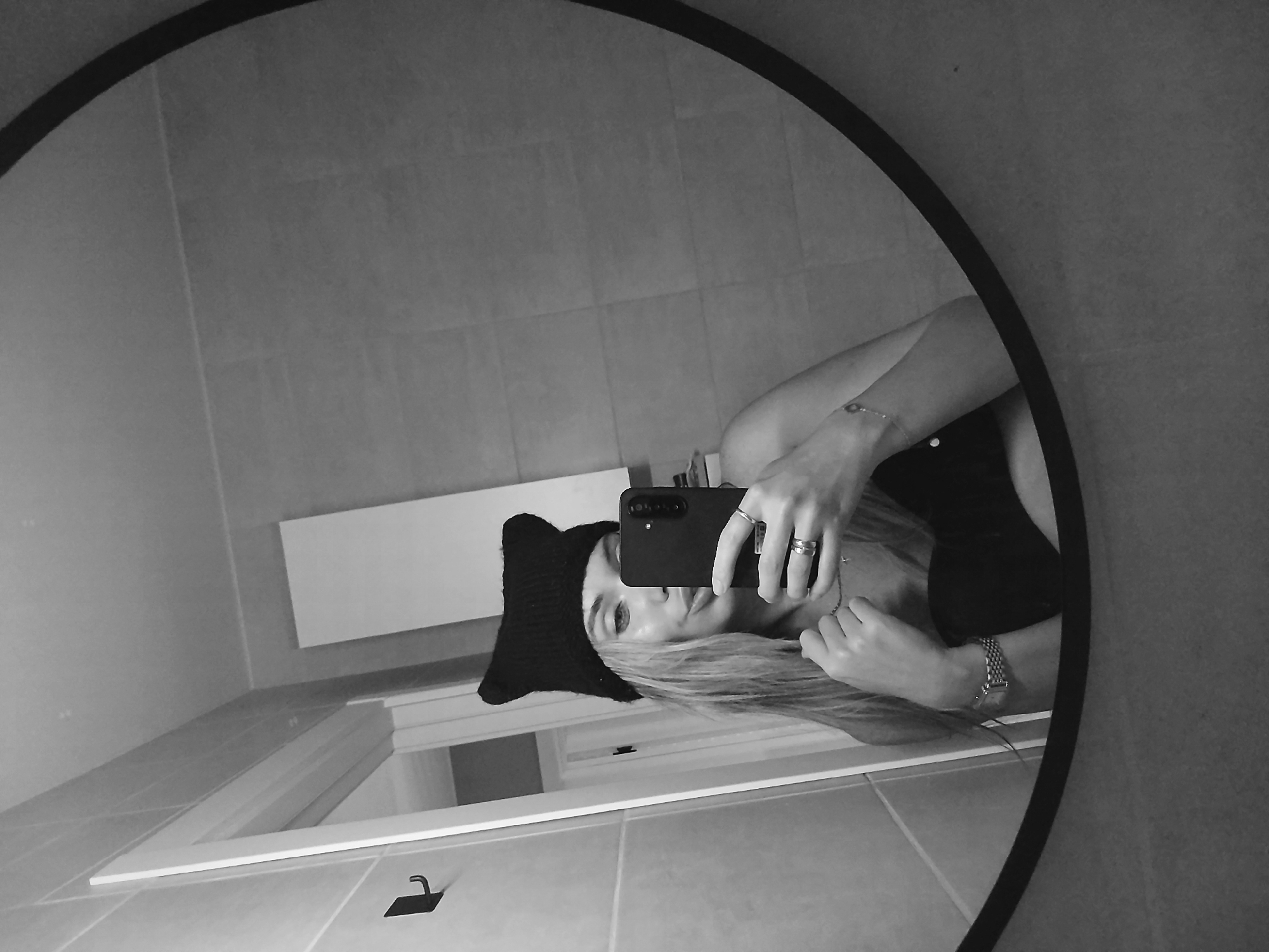 A woman is taking a selfie in a bathroom mirror. She is wearing a black hat with cat ears. Her blonde hair is visible. She is holding a black smartphone. She has rings on her fingers and a bracelet on her wrist. The bathroom has tiled walls and a vanity with a white countertop. The location is unknown and cannot be determined.