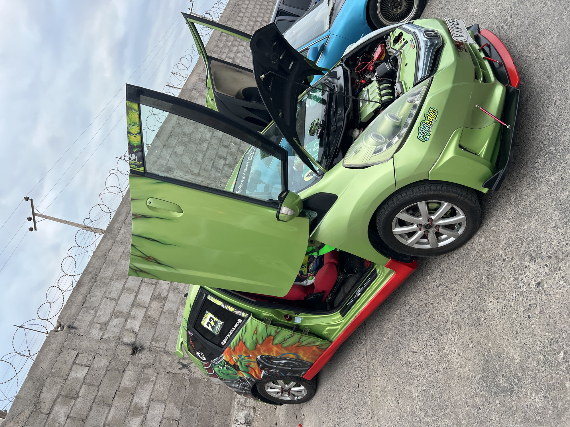 In Arica, Chile, a custom green car with its doors and hood open is parked on a concrete surface. The car features a vibrant mural on its side depicting a cartoon character with flames. The interior of the car has red racing seats. In the background, a grey concrete wall topped with barbed wire separates the parking area from an overcast sky. Another vehicle, a blue car, is partially visible to the right.