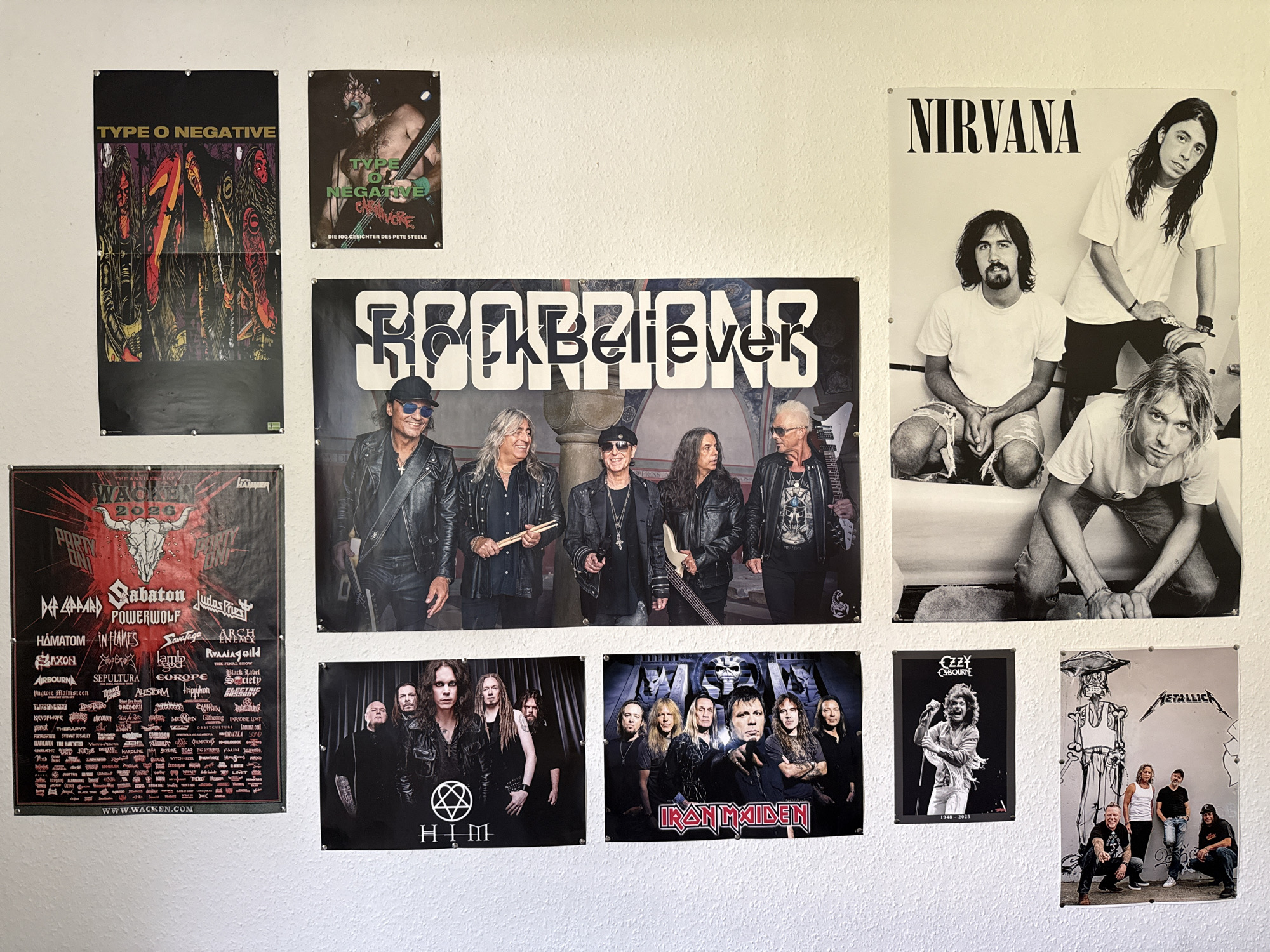 The image displays a wall adorned with various band posters, suggesting a display of musical interests. The posters depict bands such as Type O Negative, Scorpions, Nirvana, Wacken, HIM, Iron Maiden, Ozzy Osbourne, and Metallica. The wall itself appears to be painted a light, textured color, possibly white or off-white, and has a slight sheen. Several posters are pinned to the wall with metal thumbtacks, indicating they are temporarily affixed. The arrangement of the posters is somewhat eclectic, with larger ones dominating the upper sections and smaller ones filling in the lower areas.
Specifically, on the left side, a large poster of Type O Negative is prominently displayed. To its right, a smaller poster of Type O Negative is visible. Below these, a large Wacken poster is present, listing numerous bands. In the center, a large Scorpions poster is mounted. To the right of the Scorpions poster, a Nirvana poster is displayed. Below the Scorpions poster, a poster of HIM is visible, and to its right, an Iron Maiden poster. Further to the right, a smaller poster of Ozzy Osbourne is shown, and to its right, a poster of Metallica. The overall impression is of a collection of rock and metal music memorabilia.
The image was taken in the city of Bad Sachsa, Germany. This specific scene is located in a room within a building. The lighting appears to be ambient, possibly from an overhead light source or natural light entering from a window not visible in the frame. The background is consistent with an interior wall.