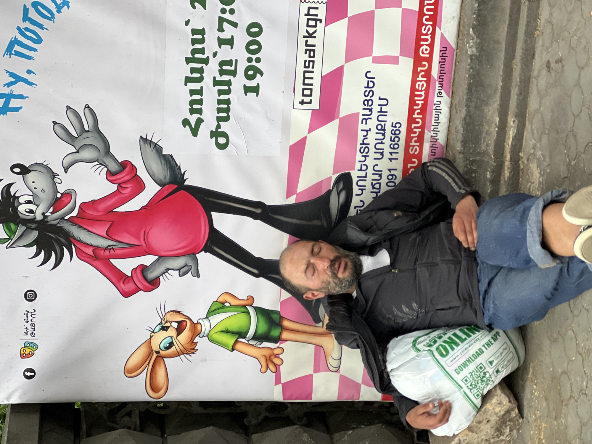 A man sleeps on the sidewalk in front of a poster in Yerevan, Armenia. The poster depicts characters from the Soviet-era cartoon "Nu, Pogodi!" advertising a performance on June 21 at 17:00 and 19:00 at the Yerevan State Puppet Theater. The poster includes the text "Ari gnanq" (Let's go) and the website "tomsarkgh." The man is lying on a plastic bag with the word "Online" and "www.yerevan-city.am" on it. The bag advertises downloading an app via Google Play and iStore. The sidewalk is cracked and uneven.