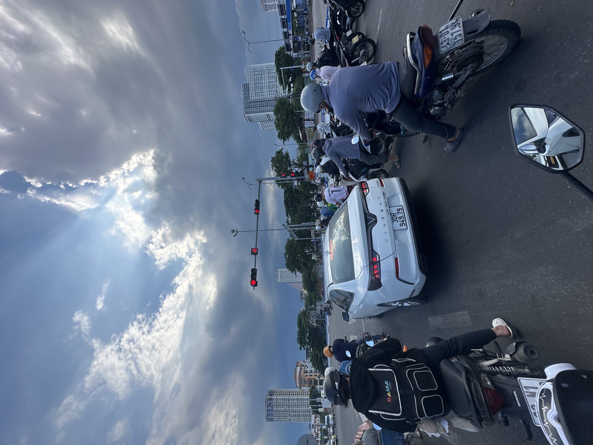 The image depicts a traffic intersection in the city of Da Nang, Vietnam, with a view towards the sky. In the foreground, several motorcycles are stopped at a red traffic light, indicating a pause in traffic flow. A white VinFast sedan is positioned in the center of the intersection, also stopped. The sky above is dramatic, with a mix of dark, stormy clouds and patches of bright sunlight, suggesting a dynamic weather condition. Tall buildings, characteristic of an urban environment, are visible in the background, framing the scene. The overall impression is one of urban life in motion, with the natural elements of the sky adding a touch of visual drama.