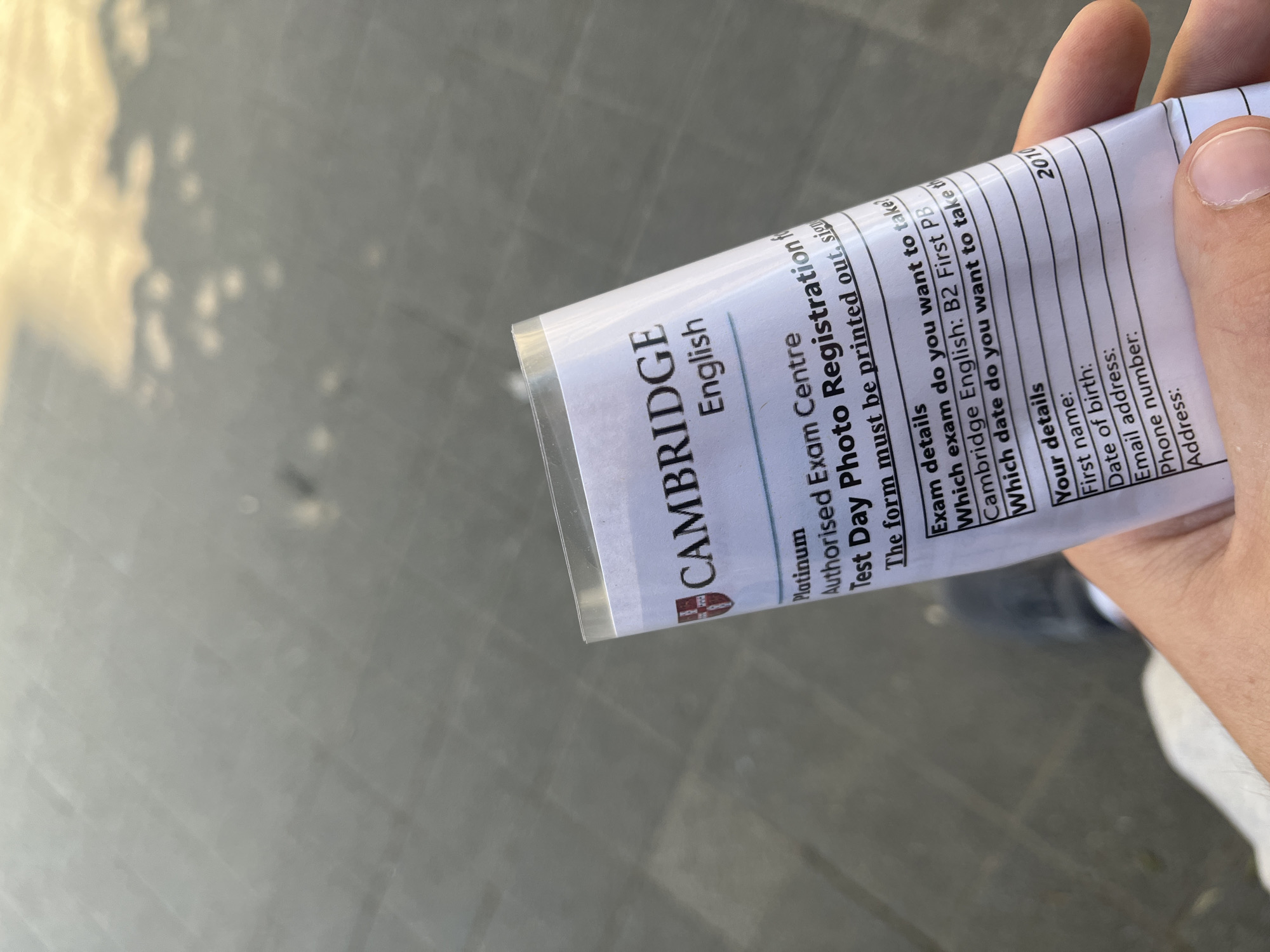 A close-up, eye-level, outdoor shot shows a hand holding a crumpled form printed with the "CAMBRIDGE English" logo. The form is a "Platinum Authorised Exam Centre Test Day Photo Registration." Below this, the form asks for "Exam details" and "Your details," with blank lines for "First name," "Date of birth," "Email address," "Phone number," and "Address." A portion of a person's jeans and shoe are visible in the lower-left corner, suggesting they are standing on a paved surface that reflects light and appears to be a street or sidewalk. The background is out of focus, showing a gray, tiled ground with reflections of light and possibly clouds in the sky. The lighting suggests it is daytime, with natural light casting shadows. The location is not identifiable as Sofia, Bulgaria.