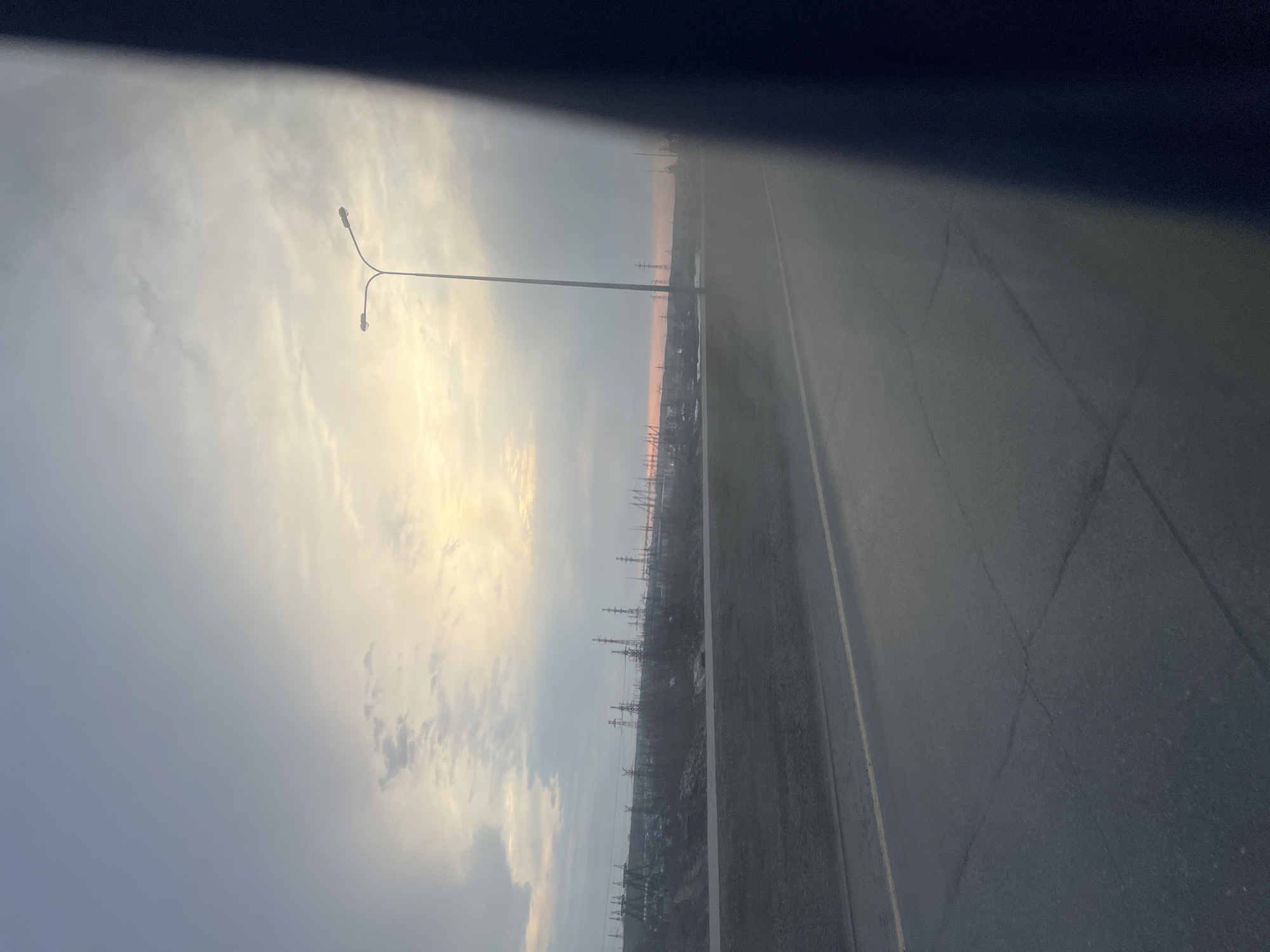 The image depicts a view from a moving vehicle, possibly on a highway or a large city road in Chaikovsky, Russia.
The road stretches horizontally across the lower right portion of the frame. The asphalt surface is gray, with visible road markings.
To the left of the road, there is an industrial area with multiple electrical pylons and structures.
The sky occupies most of the upper left of the image, exhibiting cloudy conditions with hints of a pale orange glow near the horizon. This suggests a sunrise or sunset. A tall lamppost is visible against the sky, with its dual lights near the top.
The upper part of the frame is obscured, giving the impression of looking through a car window.