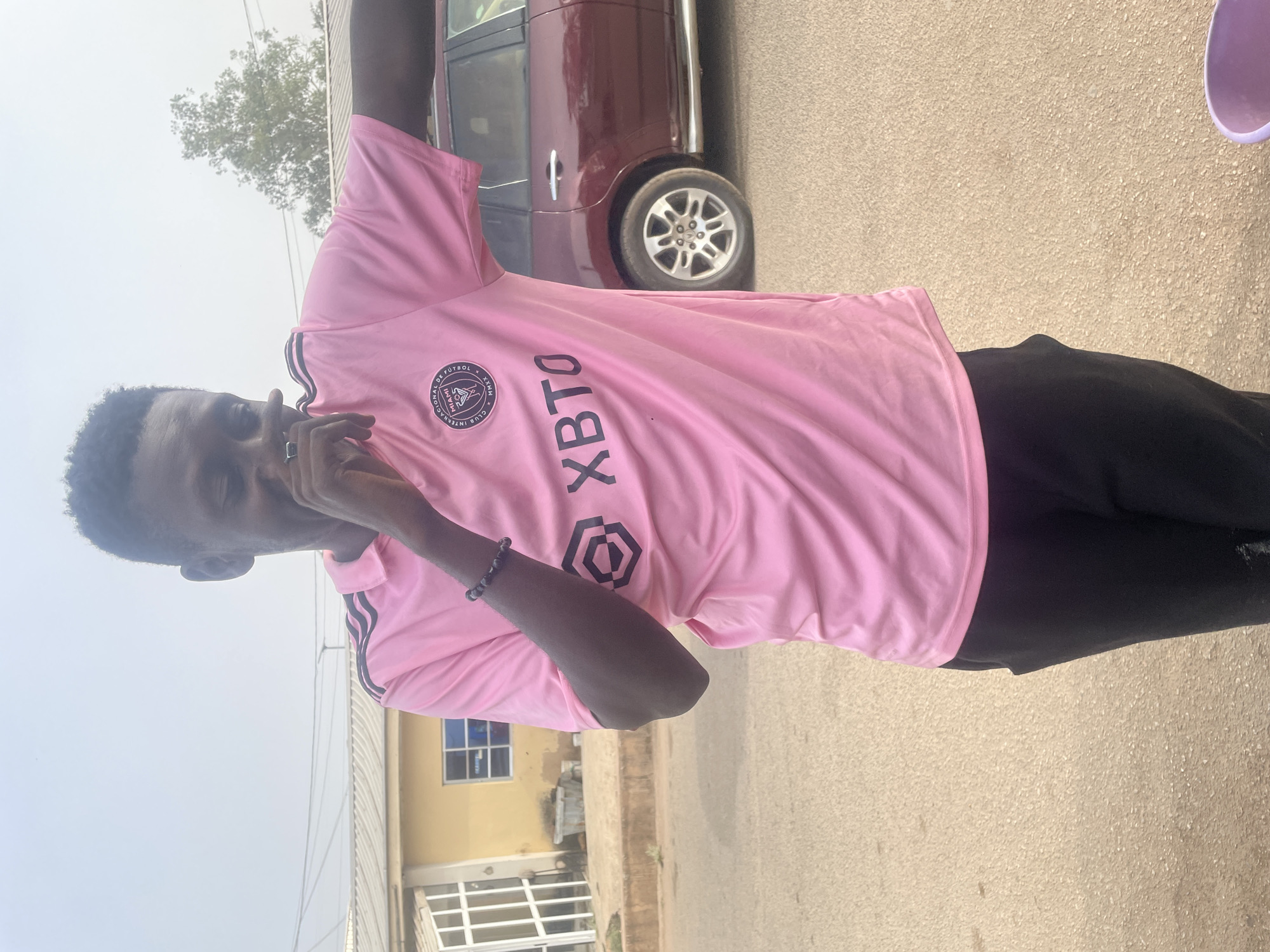 A young dark-skinned male is captured in a vertical, outdoor shot, standing under bright daylight. He is wearing a vibrant pink short-sleeved t-shirt with black accents and black trousers. The t-shirt features a circular emblem on the left chest, which reads "CLUB INTERNACIONAL DE FÚTBOL MIAMI" and "XXVII", indicative of the Inter Miami CF football club. Below the emblem, large black letters spell "XBTO". Black stripes are visible on the sleeves, suggesting an Adidas brand. He also wears a dark beaded bracelet on his right wrist and a silver ring on his left hand.
The individual is looking towards the camera with a neutral expression, his left hand raised towards his face, obscuring part of his mouth and chin.
The background shows an outdoor setting, possibly a street or a parking area in Jos, Nigeria. To the right, a dark red SUV is parked. Behind the subject on the left, a light-colored building with windows is partially visible. Above the building, a hazy, bright sky and some green tree foliage can be seen. The ground is a light-colored, textured surface, likely asphalt or concrete. The overall impression is a casual pose on a sunny day.