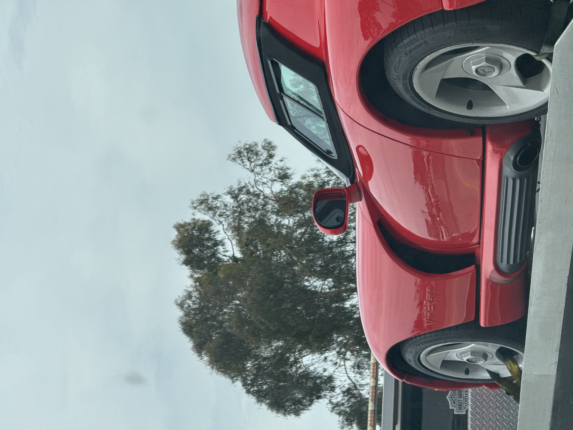 The image shows a red sports car, a Dodge Viper RT/10, partially visible on a trailer, likely in transit. The car's front wheel, side mirror, and the "Viper RT/10" emblem are clearly visible. The backdrop features a tree and an overcast sky. A portion of the trailer is visible, along with a "Harley-Davidson Motorcycles" sticker. The scene appears to be outdoors in an urban environment, however, the exact location in the city of Los Angeles cannot be determined based on the visual information provided.
