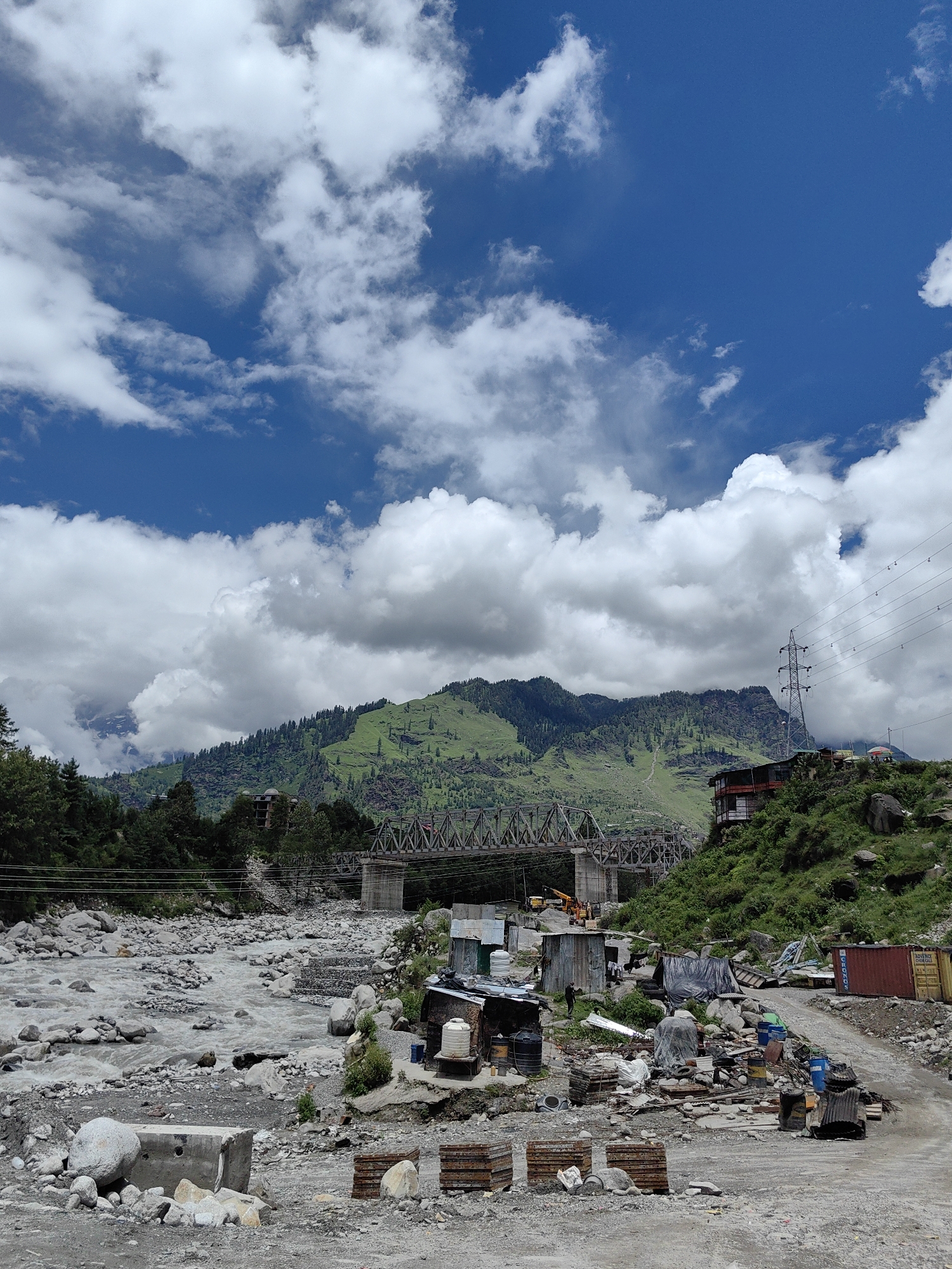 The image depicts a mountainous landscape near Kulang, India, with a river flowing through a rocky bed in the foreground. A metal truss bridge spans the river in the mid-ground, connecting two sides of the valley. On the right bank, a hillside is covered with lush green vegetation and trees, with a building situated on the higher slope. A power transmission tower and electrical lines are visible on the hill. The left bank of the river shows a rocky terrain with scattered debris, temporary structures, and construction materials, suggesting ongoing work or a settlement. The sky is a vibrant blue with scattered white clouds, indicating a clear day. The overall scene is a mix of natural beauty and signs of human activity and development in a rugged environment.