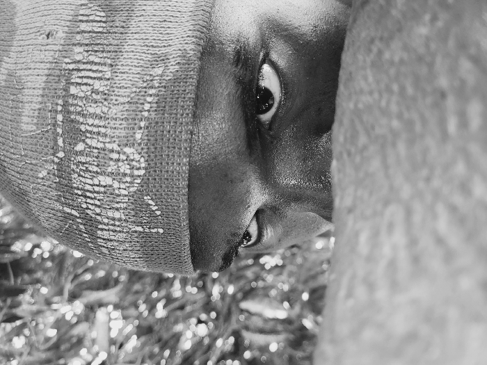 A black and white close-up photograph captures the upper face of an individual in Udu, Nigeria. The subject, exhibiting dark skin with a noticeable sheen on the forehead and around the eyes, wears a dark knitted beanie. The beanie features a faded, lighter-colored, irregular pattern across its visible surface. Both eyes are open, with pupils visible, and directed forward. A large, blurred, textured object, likely fabric or a body part, occupies the foreground, obscuring the individual's nose and mouth. The background is heavily out of focus, displaying numerous circular bokeh highlights against a darker, indistinct pattern, consistent with illuminated foliage. The individual appears to be looking over the foreground obstruction.