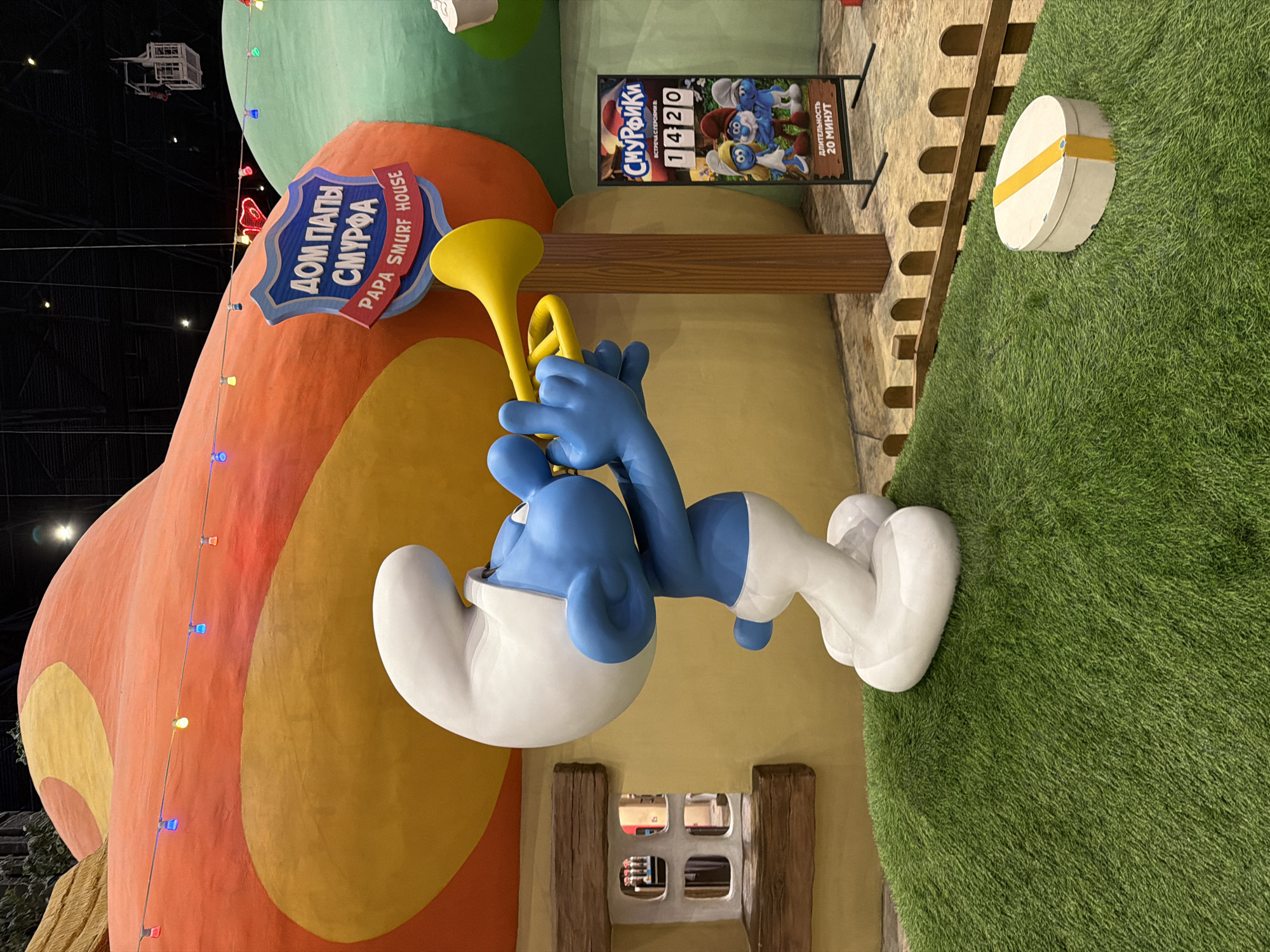 An artificial grass-covered mound in the city of Vidnoye, Russia, serves as a stage for a large statue of a Smurf blowing a yellow trumpet. The Smurf, depicted in its characteristic blue skin, white hat, and white pants, stands on its hind legs, its front paws holding the instrument to its lips. In the background, a colorful, oversized mushroom-shaped structure dominates the scene, painted in shades of orange and yellow, with a sign that reads "ДОМ ПАПЫ СМУРФА PAPA SMURF HOUSE" in blue and red lettering. String lights with colored bulbs are draped across the mushroom structure. To the right of the Smurf statue, a wooden post supports a vertical sign displaying "СМУРФИКИ" (Smurfs) and an image of several Smurfs, along with text indicating a meeting with characters and a duration of 20 minutes. A low, wooden fence borders the grass mound. On the grass to the right of the Smurf statue, a white oval-shaped object with a yellow band is placed. The environment appears to be an indoor entertainment space, possibly a theme park or children's attraction, with dark ceilings and artificial lighting visible.