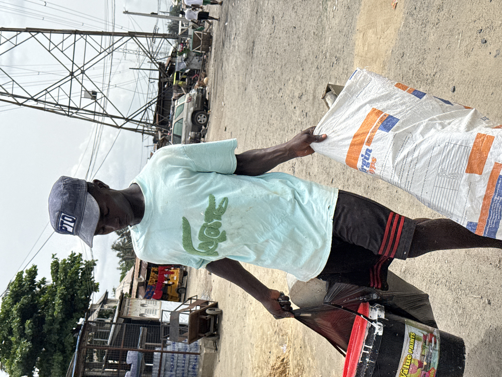 A medium shot, taken outdoors in the city of Ado, Nigeria, shows a man standing on a street with electrical towers in the background. He wears a blue hat with "PTO" on it, a light blue Lacoste t-shirt, and black shorts with red stripes. He holds a large white bag labeled "Virgin" in his right hand and a black trash bag with a black paint bucket inside in his left. In the background are various buildings, vehicles, and objects lining the street. The ground is a light brown, sandy color.