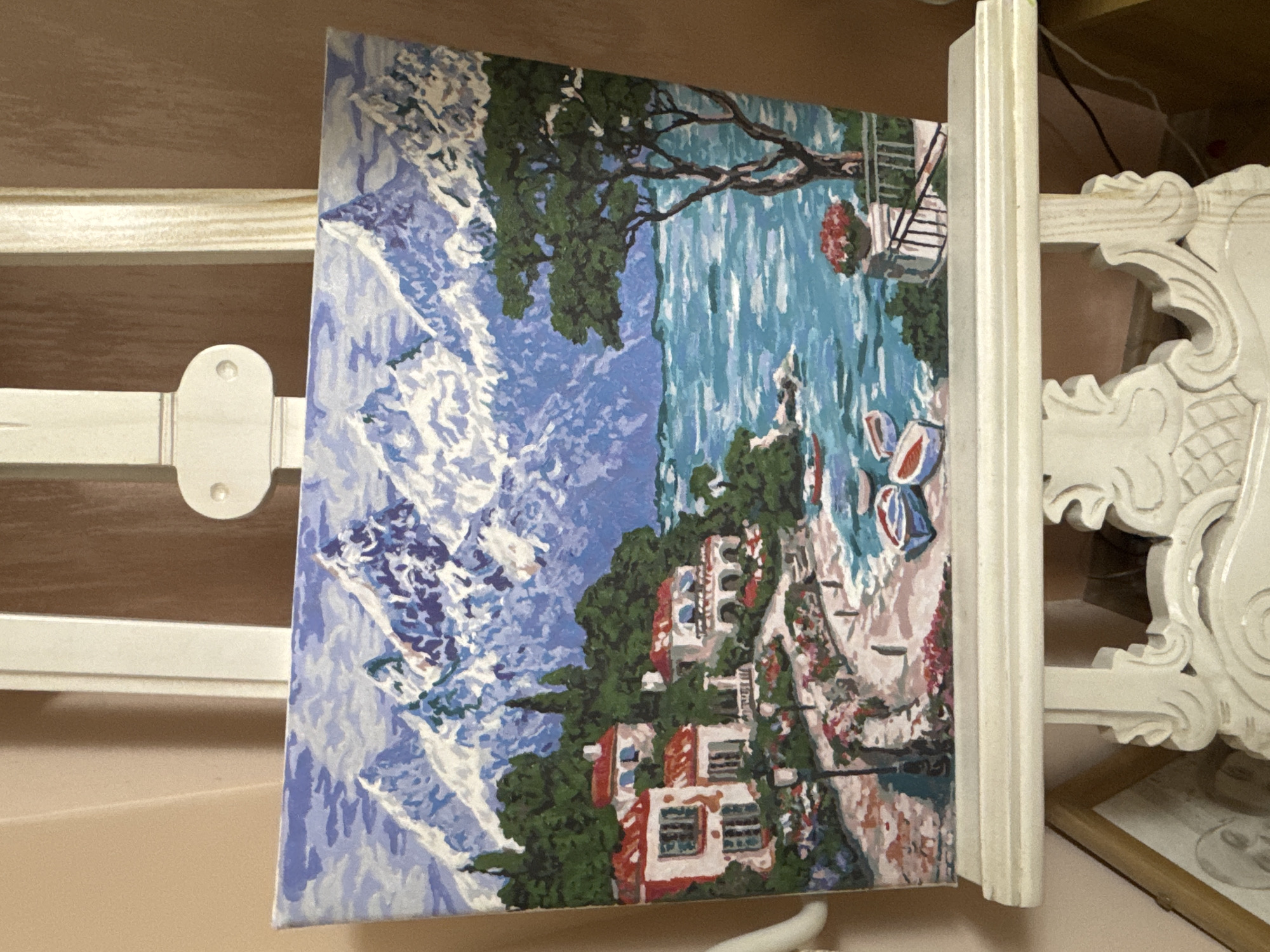 A painting depicting a coastal scene is propped up on a white easel. The painting portrays a coastline with houses nestled among trees, blue water with boats, and a rocky shore. The style of the painting is somewhat impressionistic. The objects surrounding the easel cannot be identified. The location depicted in the painting is not in the city of Plovdiv, Bulgaria.