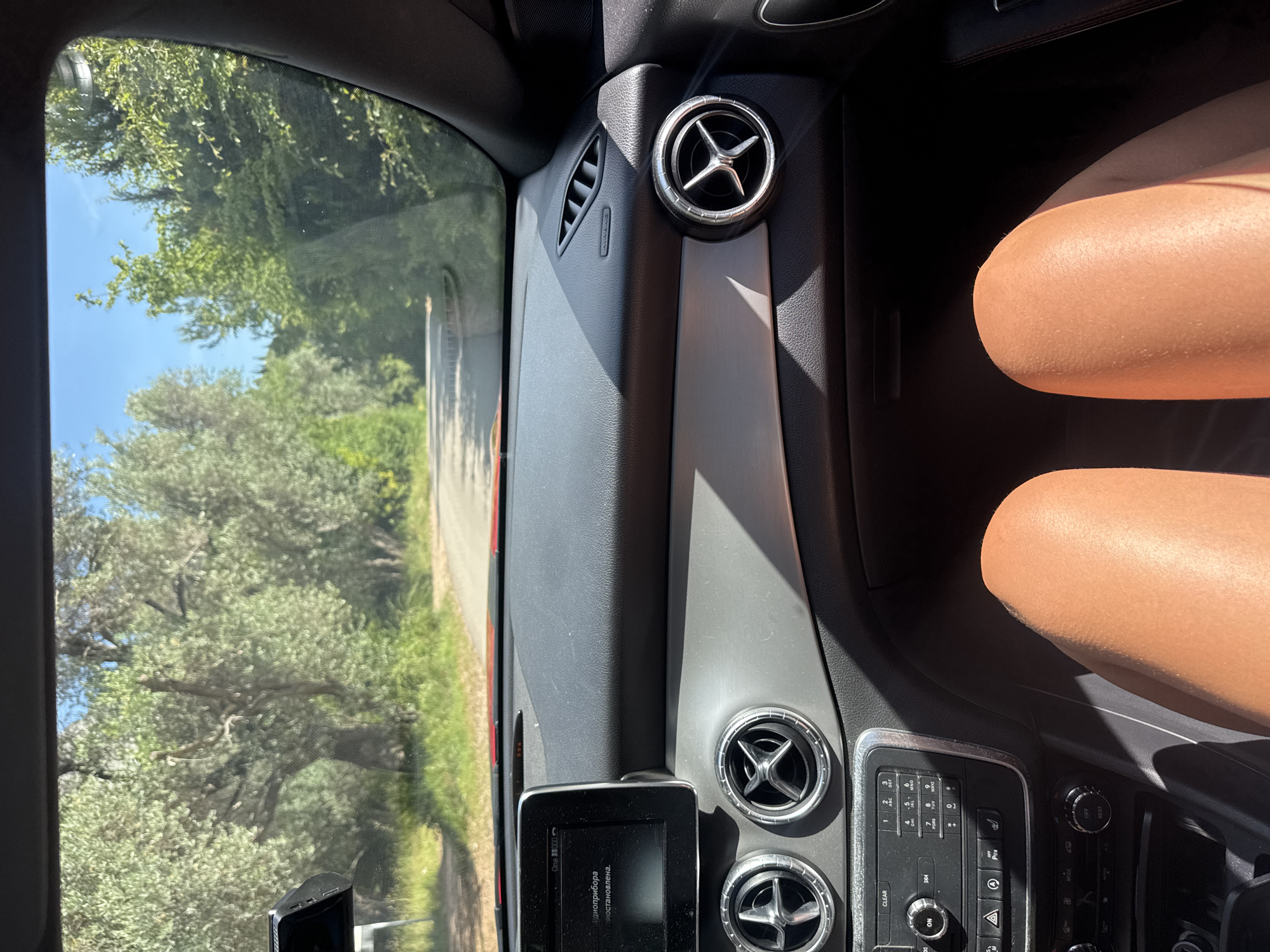 The image depicts a first-person perspective from the passenger seat of a car, looking forward through the windshield. The lower portion of the frame shows the bare, tanned thighs and knees of a person, illuminated by sunlight. The car's interior features a dark grey dashboard with a red trim line visible along the base of the windshield. A silver accent strip runs horizontally across the passenger side of the dashboard. Three circular air vents with chrome rings and star-shaped grilles are visible across the dash, with a fourth partially visible on the far right. An infotainment screen on the driver's side displays text, partially visible as "One 000" and what appears to be Cyrillic script below it. Below the screen are car controls including a power/volume knob, media buttons, a numeric keypad, hazard light button, and climate controls. An "AIRBAG" label and a small vent are visible on the passenger side A-pillar area. Sunlight creates strong shadows within the car's interior.
Outside the car, a narrow, paved road is visible, curving gently to the right. The road is flanked by grassy verges and dense vegetation. Numerous trees, appearing to be olive trees with their characteristic light green foliage, line both sides of the road. Beyond the trees, a clear, bright blue sky is visible. The scene suggests a sunny day in a natural, tree-lined environment. The location is specified as Sveti Stefan, Montenegro.