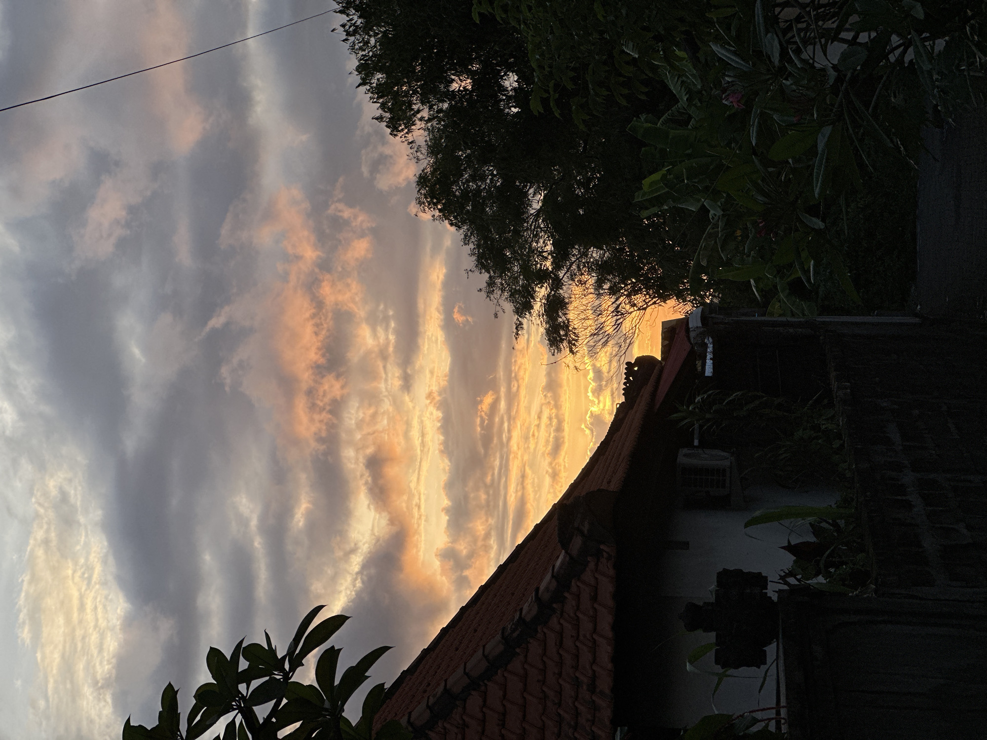 The image shows a sunset sky above the city of Dalung, Indonesia. The sky contains layers of gray and orange clouds. A dark tree and the roof of a building are silhouetted in the foreground. There is an electrical wire running across the top left corner of the image. The lighting suggests that it is near sunset.
