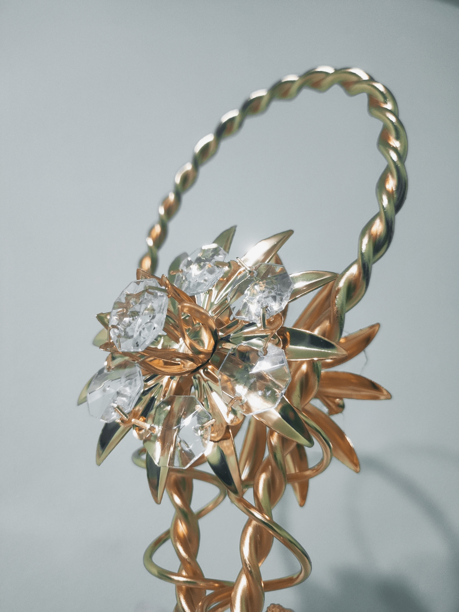 This is a close-up, vertical shot of a decorative, golden object. The object features a twisted, looped handle that leads down to a floral embellishment. This embellishment consists of metallic, petal-like shapes and several clear, faceted crystals that resemble gemstones. The metallic parts are a bright, polished gold, reflecting the light. The crystals are also highly reflective, scattering light and creating bright highlights. The background is a plain, light gray, which is out of focus, drawing all attention to the object. The lighting appears soft and diffused, casting gentle shadows on the background. There is no text visible in the image.