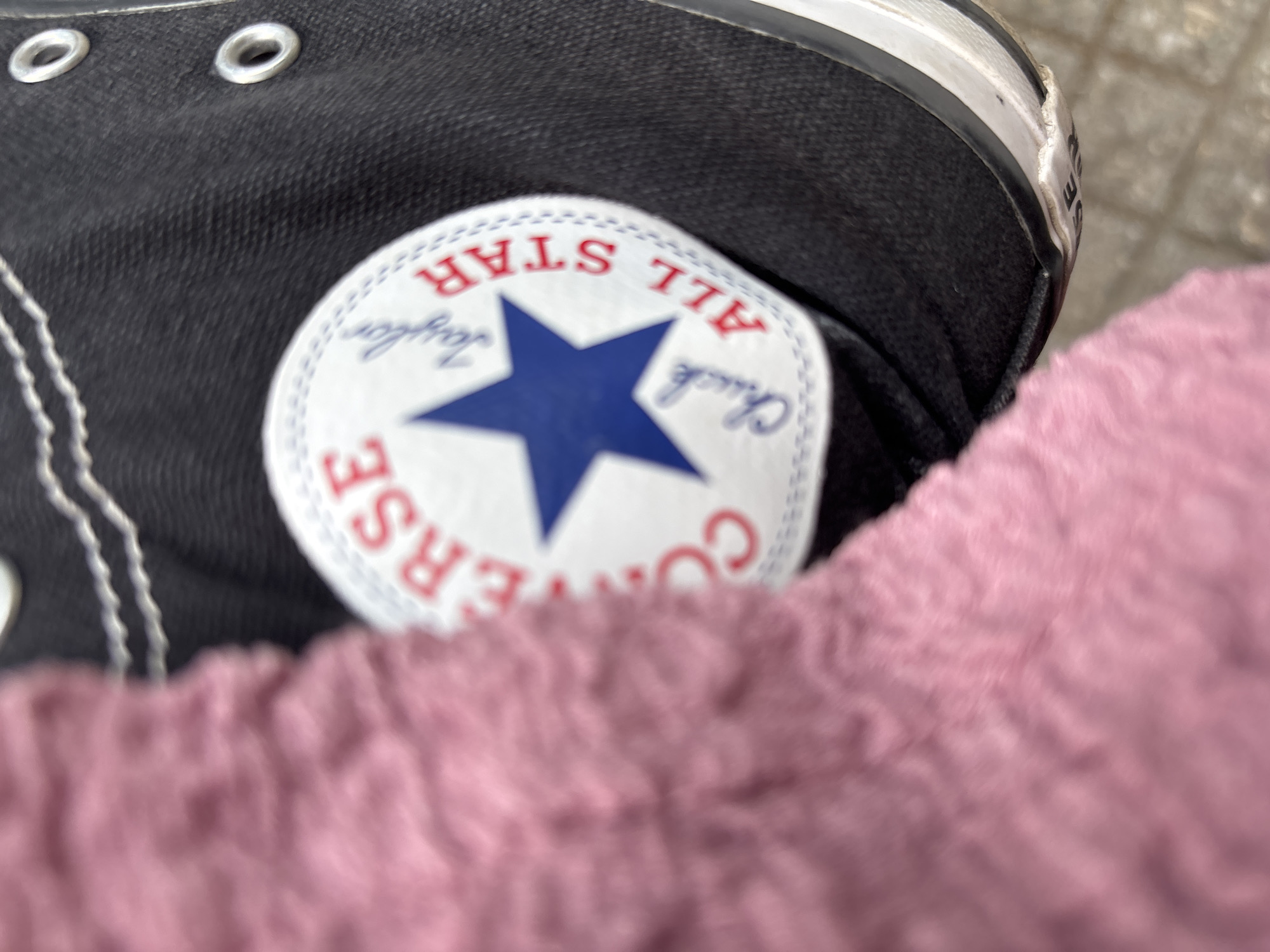 This is a close-up, angled shot of a black Converse All Star sneaker. The shoe is partially covered by a soft, pink fuzzy material, possibly clothing. The iconic circular Converse logo is visible on the side of the shoe, featuring a blue star and text that reads "CONVERSE ALL STAR" and "Chuck Taylor." The stitching on the shoe is white. The background is blurred and appears to be an outdoor setting with a textured, light-colored surface, possibly pavement or cobblestones, and a hint of a vertical object. The lighting suggests it could be daytime. The image is focused on the detail of the shoe and logo. No people are explicitly visible, but the presence of the shoe implies a person is wearing it. The location context is Gotse Delchev, Bulgaria.