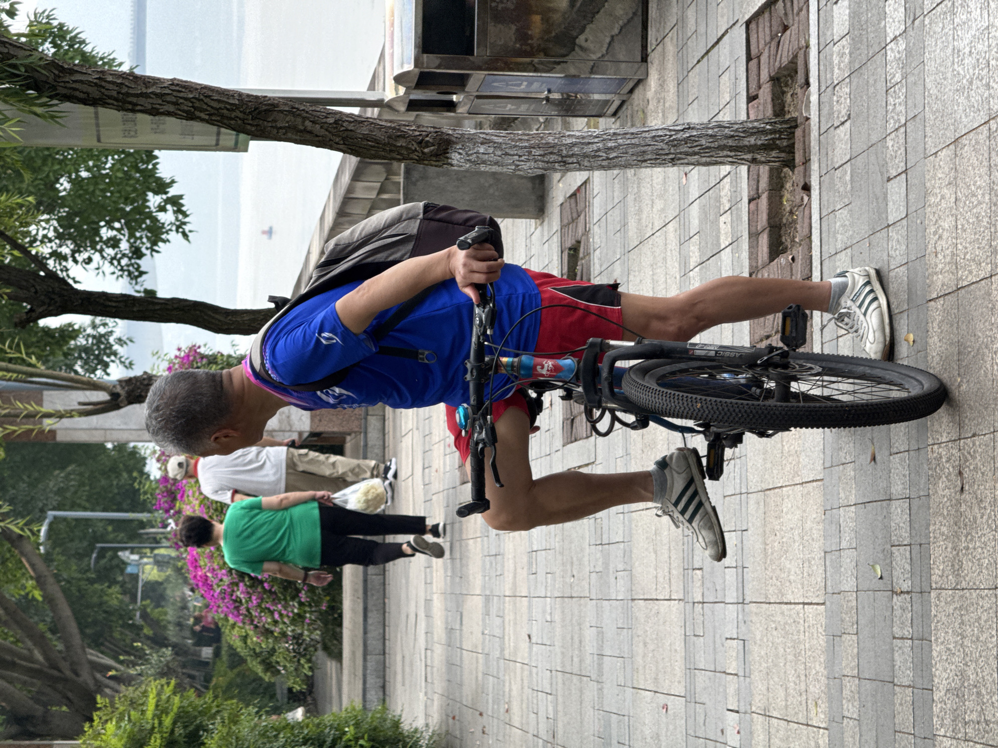 A male individual stands beside a bicycle on a grey paver sidewalk in Jiangjin, China, adjacent to a large body of water. The man, with short grey hair, wears a blue short-sleeved athletic shirt with a white logo, red athletic shorts, grey socks, and white athletic shoes with three dark stripes. A grey backpack is on his shoulders, and a pink lanyard is visible around his neck. His right foot rests on the left pedal of the dark-colored mountain bike-style bicycle, and his left foot is on the ground. His right hand grips the bicycle's handlebar.
In the mid-ground, two individuals walk away from the camera on the same sidewalk. The leading person is a woman with dark hair, wearing a green t-shirt and black pants, carrying a translucent plastic bag containing light-colored items in her left hand. Behind her, a man wearing a light-colored baseball cap, a white polo shirt with a red collar, and khaki pants follows.
The sidewalk is bordered on the left by lush green foliage and dense bushes with vibrant magenta flowers. In the background, several trees with visible trunks and green canopies are present. A concrete retaining wall separates the sidewalk from the wide body of water, which appears calm and light grey under an overcast sky. A small, red, conical buoy or marker is visible on the water's surface, with a distant, hazy landmass on the horizon. Two metallic trash receptacles are positioned on the right side of the sidewalk, next to a tree with a white-painted base.