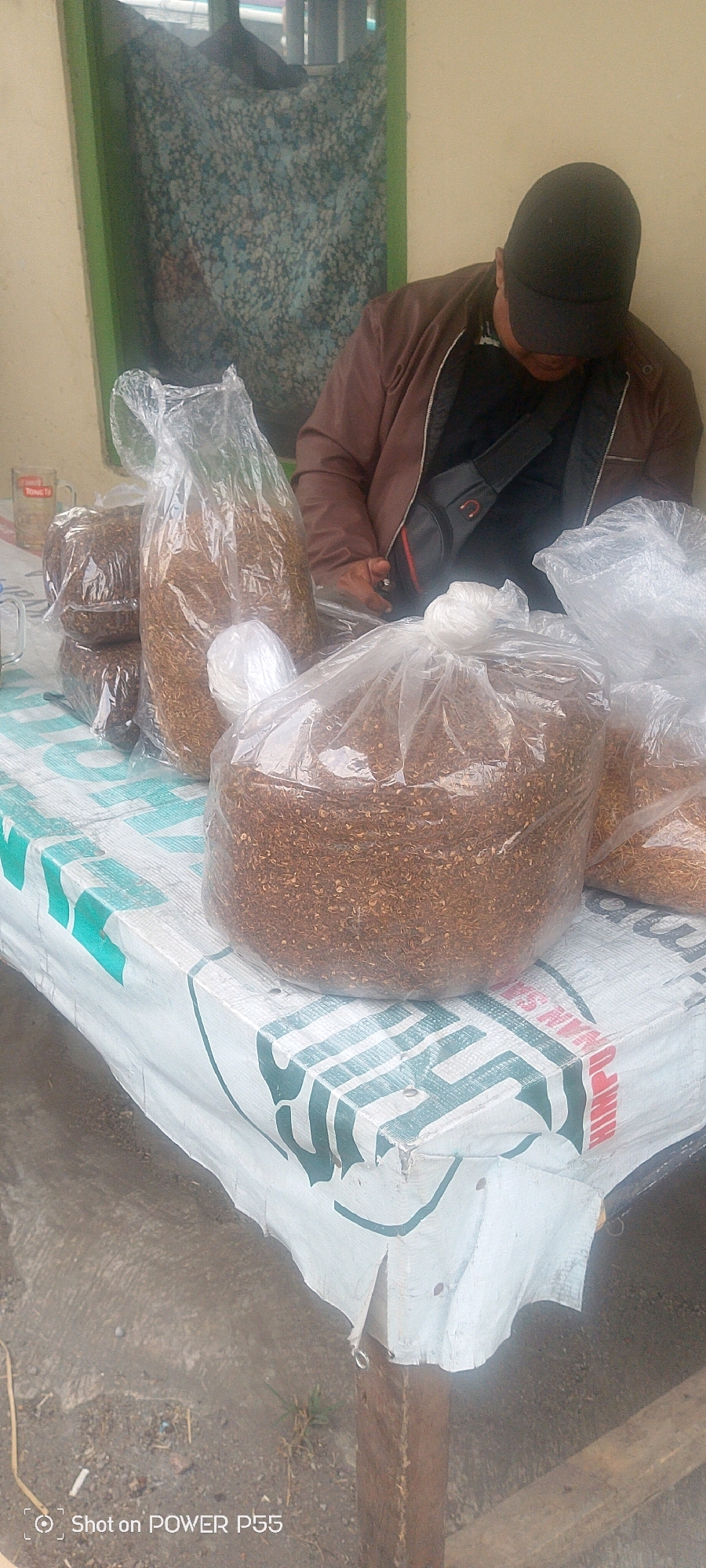 A person wearing a dark baseball cap and a brown jacket is seated behind a table covered with a white and green patterned tarp. The person is looking down at something on the table. Several large, clear plastic bags filled with a brown, granular substance, appearing to be dried tobacco, are displayed on the table. Some of the bags are stacked, and one is partially obscured by another. In the background, a window with a floral patterned curtain is visible. A glass mug with red lettering is also present on the table. The setting appears to be outdoors or in an open-air market. The bottom right corner of the image contains a watermark that reads "Shot on POWER P55".