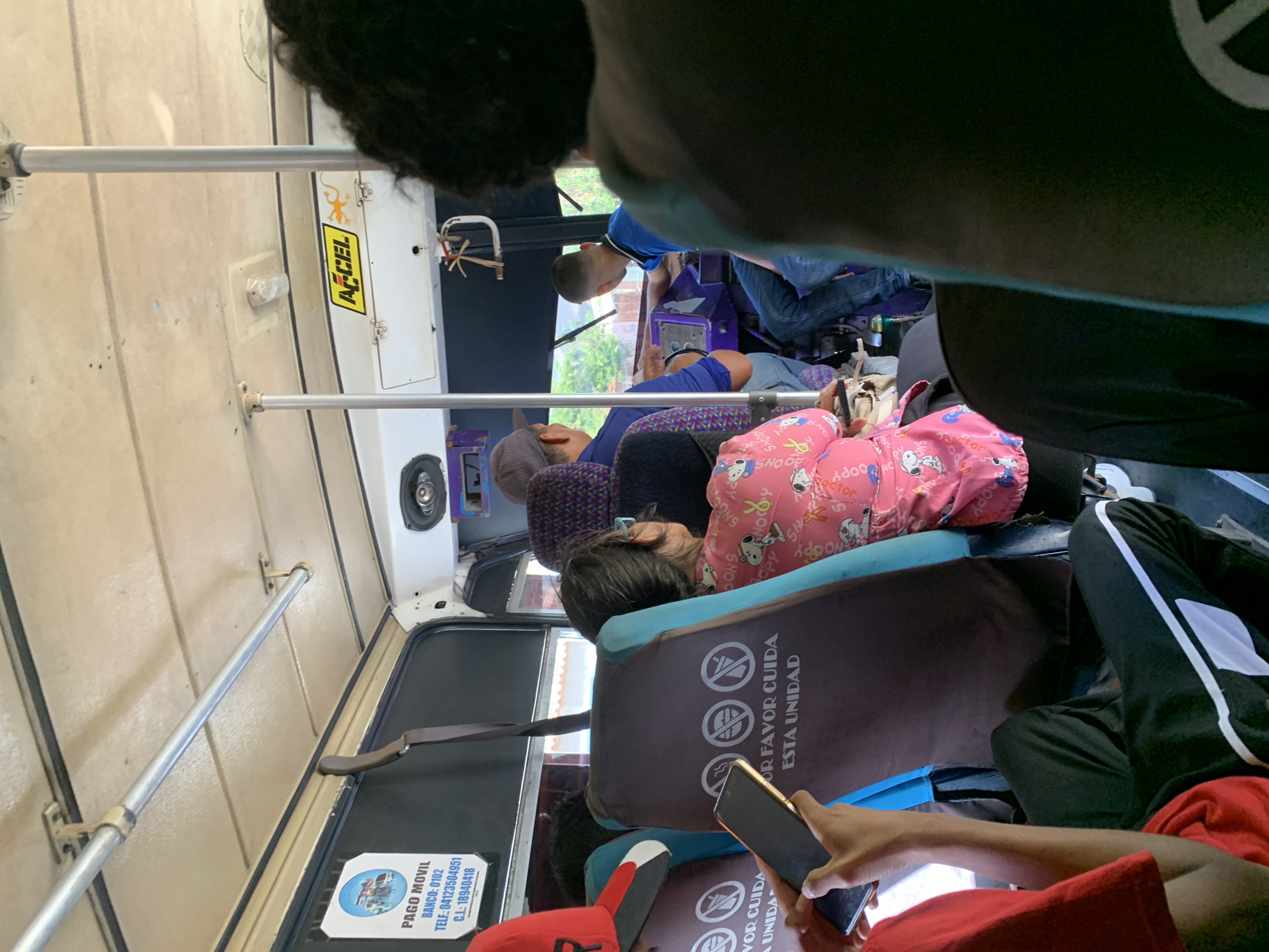 The image, taken inside a public transport bus in La Asuncion, Venezuela, captures a candid view of passengers during daytime. The perspective is from the front of the bus, looking towards the back, showing a bustling but relaxed atmosphere.
In the foreground, parts of a person's head with dark, curly hair and shoulder are visible on the upper left, likely the person holding the camera. Directly below, a passenger in a red shirt and black track pants is engrossed in a smartphone, holding it with both hands. To their left, another person in a red cap with a white "B" logo is partially visible.
Further into the bus, a child or small person is seen sleeping soundly on a seat, their head resting against the seat in front. They are wearing a bright pink top adorned with Snoopy characters and "Snoopy" text, along with other faint words like "doctor." Behind them, another passenger in a blue shirt is visible. Toward what appears to be the front of the bus (or further back in the vehicle depending on orientation), another individual with a short haircut, also in a blue shirt, looks out a window.
The bus interior features overhead grab rails, beige paneling, and speakers mounted near the ceiling. On the back of several seats, stickers with the text "POR FAVOR CUIDA ESTA UNIDAD" (Please take care of this unit) are visible, accompanied by three circular "no" symbols, likely indicating rules such as no feet on seats or no littering. A "PAGO MOVIL" (Mobile Payment) sticker is prominently displayed on a window or panel, showing bank details ("BANCO: 0102"), a telephone number ("TELF: 04123504951"), and an identification number ("C.I.: 18940418"), along with an image of a bus. Another yellow sticker with "AECEL" and a lizard logo is affixed to a white panel. The bright natural light suggests it is daytime.