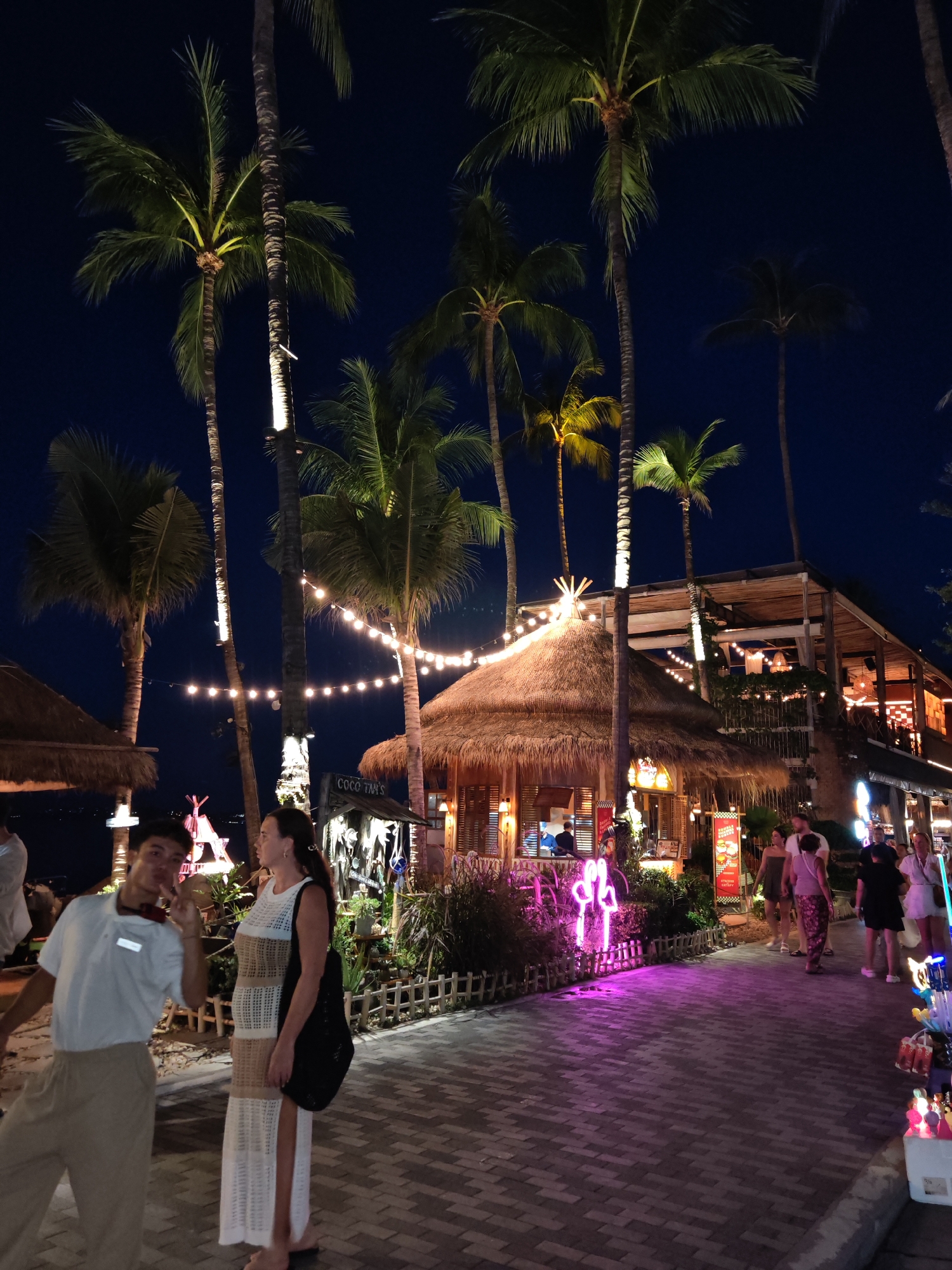 In Baan Bo Phut, Thailand, at night, a young man wearing a white polo shirt and beige pants stands on a brick pathway, making a peace sign with his right hand. Next to him, a woman in a long, crocheted dress with white and tan stripes looks to her right, carrying a black shoulder bag. Behind them, a thatched-roof hut with open sides is illuminated by string lights and neon signage. Palm trees with illuminated trunks and fronds tower over the scene. In the background, a multi-level building with wooden accents and more string lights is visible. Several other people are walking along the pathway in the distance, and various stalls selling goods are set up. The overall atmosphere is vibrant and festive, typical of a tropical beachside resort town at night.