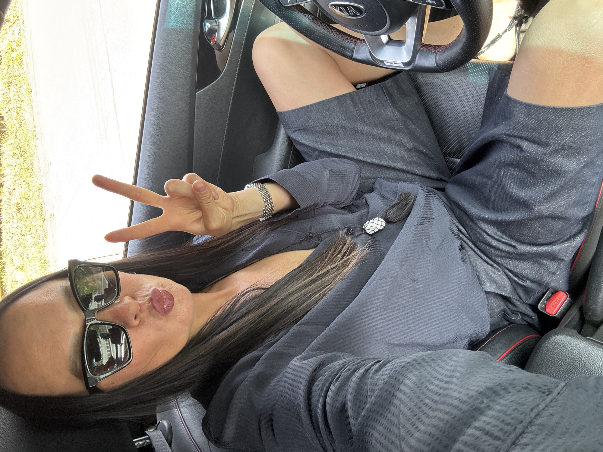 A woman is sitting in the driver's seat of a Kia vehicle, oriented horizontally in the frame. She wears sunglasses, a dark, buttoned shirt, and dark shorts. Her legs are propped up near the steering wheel. She is giving the peace sign with her left hand and has puckered lips as if posing for a selfie. The interior of the car is visible, including the dashboard and seats. The scene is sunlit, suggesting it is daytime. Outside the window, greenery is visible. It is impossible to confirm or deny the image was taken in Chaikovsky, Russia.