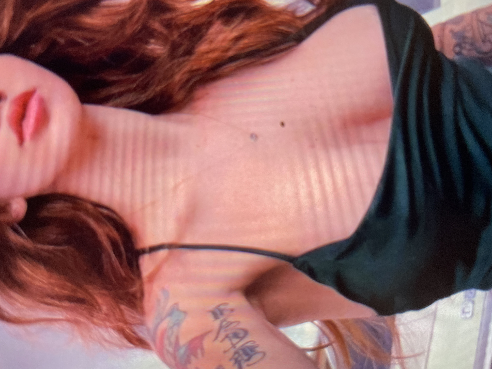 The image shows a close-up, vertically oriented, top-down view of a person's upper body, with a focus on their chest, neck, and hair. The person has fair skin with freckles, and long, reddish-brown hair that falls around their shoulders and chest. They are wearing a dark green, strappy top. Part of a tattoo is visible on their arm, featuring colorful designs and Japanese characters. The person's face is partially visible on the left side of the frame, showing their lips. The lighting suggests an indoor setting, though no specific location cues are apparent. There is no indication of the time of day or weather. The overall impression is intimate and personal, with no discernible activity or event taking place beyond the pose itself.