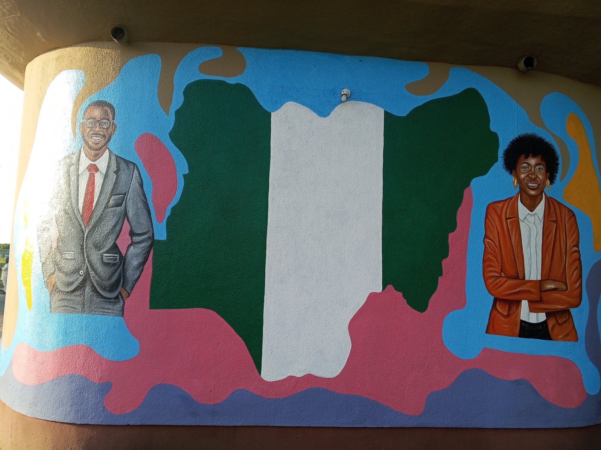 A mural in Tawpo, Nigeria, features a painted map of Nigeria in green and white. A painted portrait of a man in a gray suit and a red tie stands to the left of the map. To the right of the map is a painted portrait of a woman in an orange blazer and a white shirt. The background behind the map is painted with brown, blue, and pink colors. The mural appears to be on the exterior of a building.