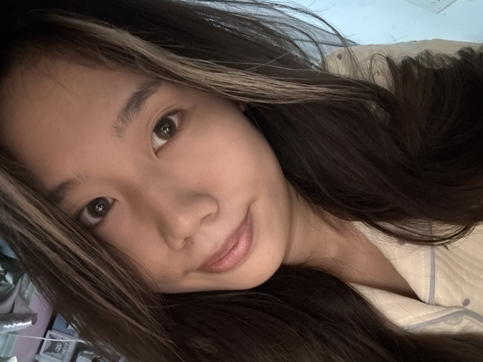The image presents a close-up, head-and-shoulders selfie of a single individual, an East Asian female, located in an indoor setting within Nan'an District, China. The subject is positioned centrally, looking directly into the camera with a slight head tilt and a subtle, closed-mouth smile. She has long, dark brown hair with lighter, blonde or grey streaks visible near the forehead and along the right side. Her eyes are dark brown. She is dressed in a light-colored, possibly cream or white, collared garment, appearing to be a quilted pajama top, accented with light blue piping along the lapel and small, light-colored buttons.
The background is out of focus and dimly lit. To the subject's left, an indistinct collection of items is visible on a surface, including a framed object that appears to depict a cartoon character resembling Sanrio's Kuromi. To the subject's right, a solid light-blue wall is partially visible, featuring a white electrical outlet or switch plate mounted on it. The ambient lighting suggests an artificial indoor environment.