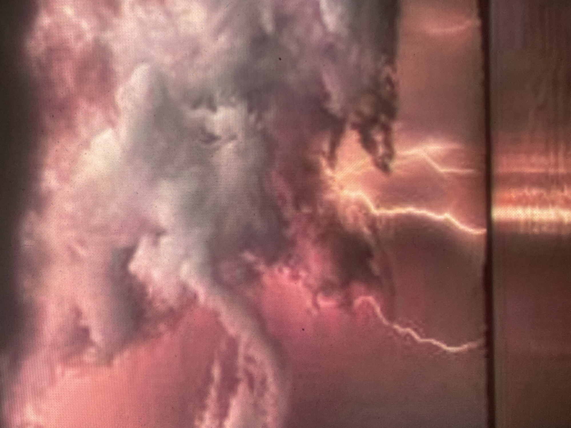 The image captures an intense electrical storm over a body of water in the city of Zelenyi Hai, Ukraine. The sky is dominated by heavy, turbulent clouds, displaying a color palette ranging from deep red and pink on the left to darker purplish-gray on the right. A pervasive reddish glow emanates from within the cloud layer. Multiple lightning strikes are visible; two prominent, brightly luminous, yellow-white bolts descend centrally towards the water's surface, flanked by a less intense, jagged bolt on the far left and a faint vertical streak on the right. The calm surface of the water reflects the reddish ambient light from the sky and the intense brilliance of the lightning. A dark, low-lying landmass or distant tree line forms a silhouetted horizon between the water and the sky. No human subjects, discernible man-made structures, or direct interactions are present within the scene, which depicts severe weather conditions.