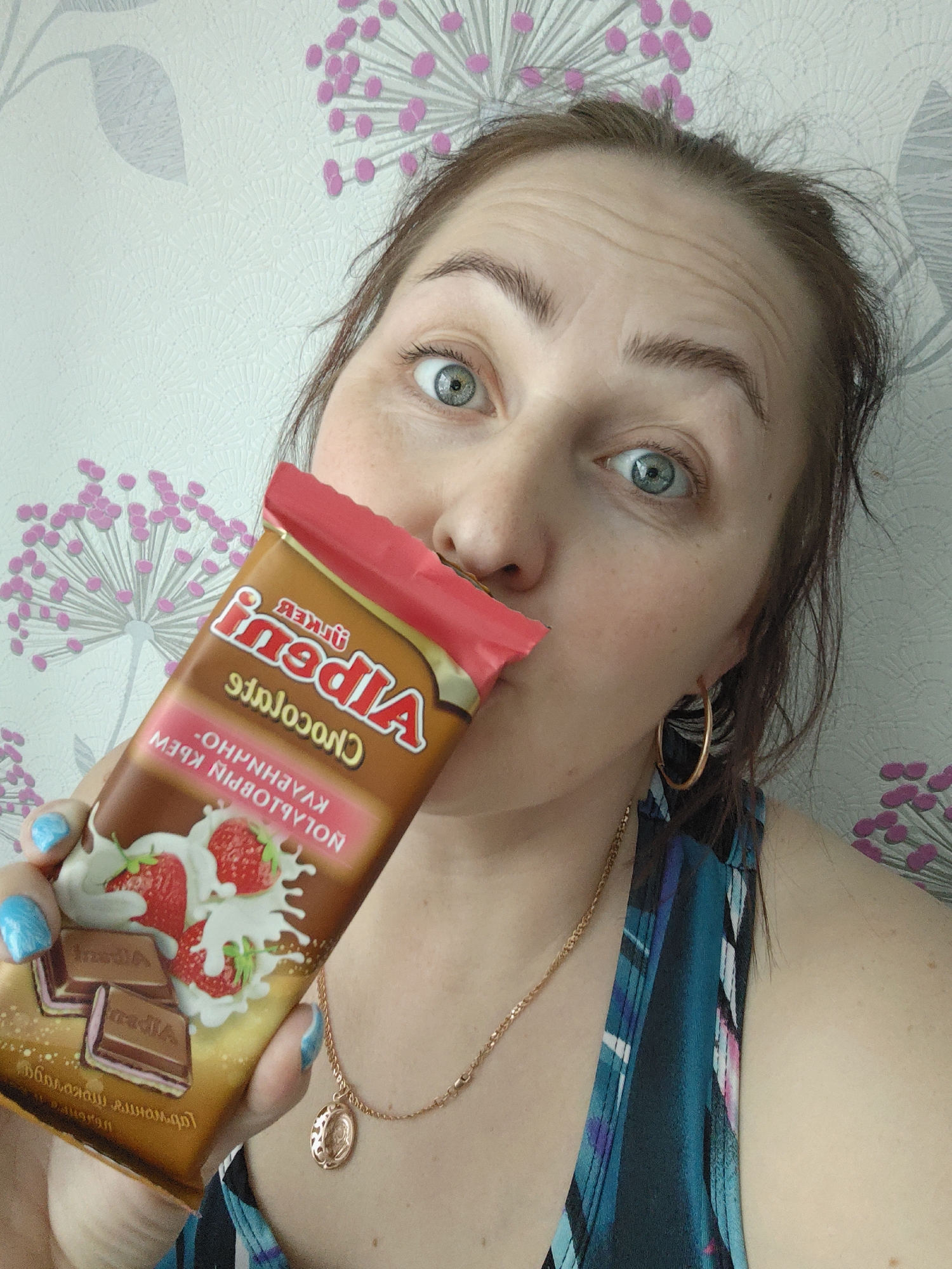 A woman is holding a chocolate bar in front of her face, partially obscuring her nose. The chocolate bar is brown with a red top and features an image of strawberries and chocolate squares. The text on the wrapper is in Cyrillic. The woman has light skin, green eyes, and brown hair pulled back. She is wearing gold hoop earrings and a gold necklace with a pendant. Her nails are painted blue with a white design. She is wearing a blue and green patterned top. The background is a wall with a white and pink floral pattern. The location is Markovskii, Russia.