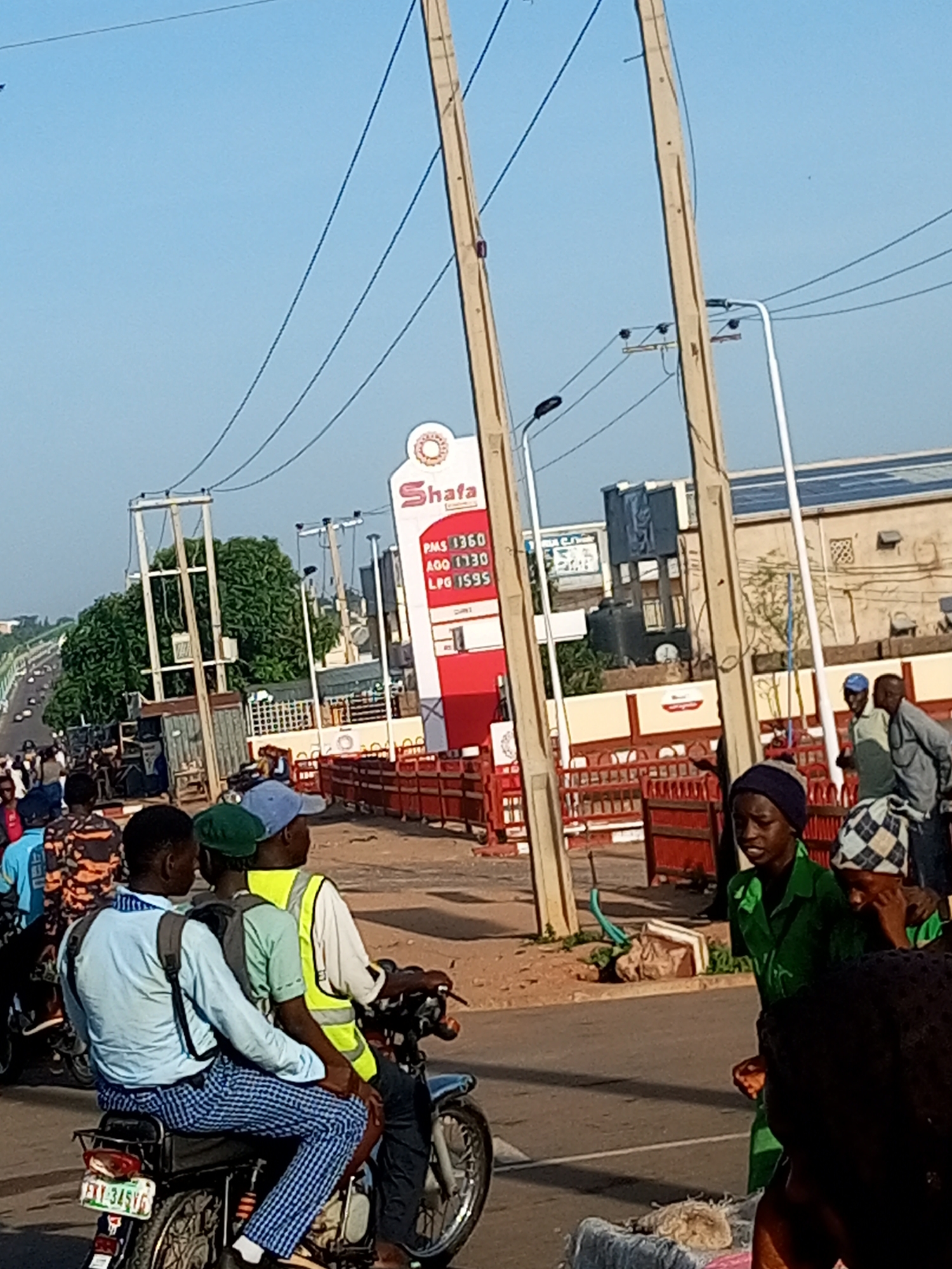 A street scene shows several people, a motorcycle, a gas station sign, and power lines. Three men are riding on a motorcycle in the foreground, with the driver wearing a light blue long-sleeved shirt and checkered pants, the middle passenger a green shirt and a blue cap, and the rear passenger a yellow vest. Behind them, a red and white gas station sign displays prices: "Shata PMS 1360 AGO 1130 LPG 1599." Several people are standing near the sign, and others are visible in the background on the street and sidewalk. Two utility poles with power lines run diagonally across the frame. The environment appears to be an urban setting with a clear blue sky.