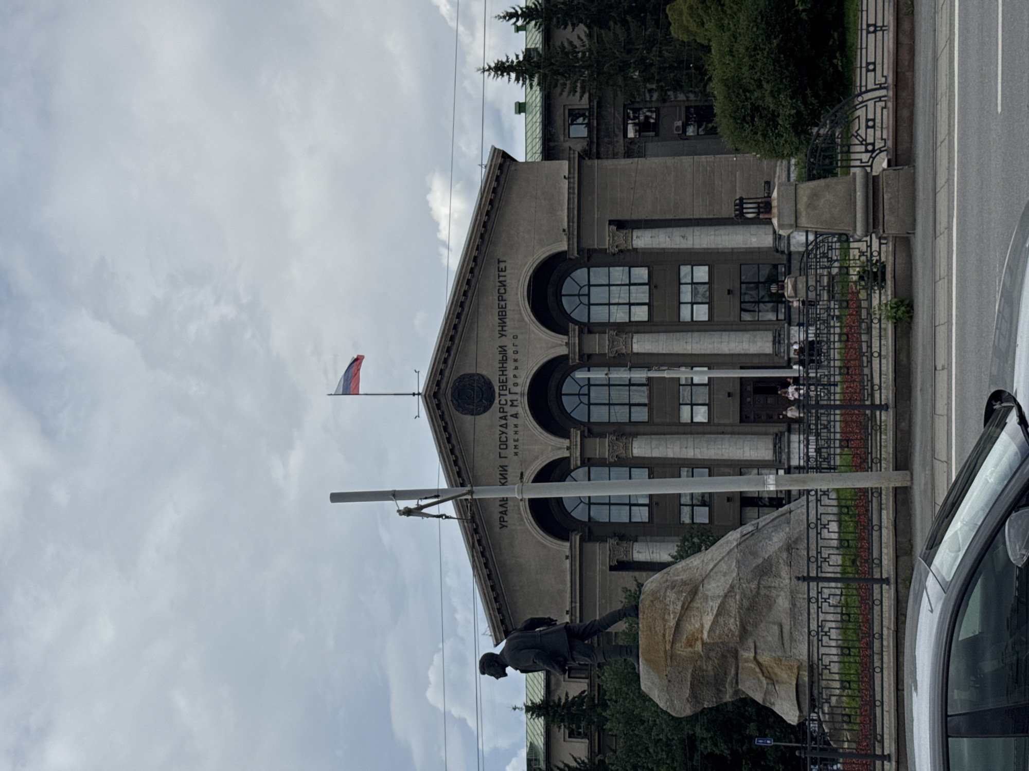 The image shows a statue of a man standing on a rock in front of a large, imposing building with the inscription "УРАЛЬСКИЙ ГОСУДАРСТВЕННЫЙ УНИВЕРСИТЕТ имени А.М. Горького" (Ural State University named after A.M. Gorky). The building has a neoclassical facade with columns and arched windows. A Russian flag is visible on top of the building. The statue is located in a public space with a fence and greenery. The scene appears to be in Yekaterinburg, Russia, as suggested by the university's name. A car is partially visible in the foreground. The sky is cloudy.