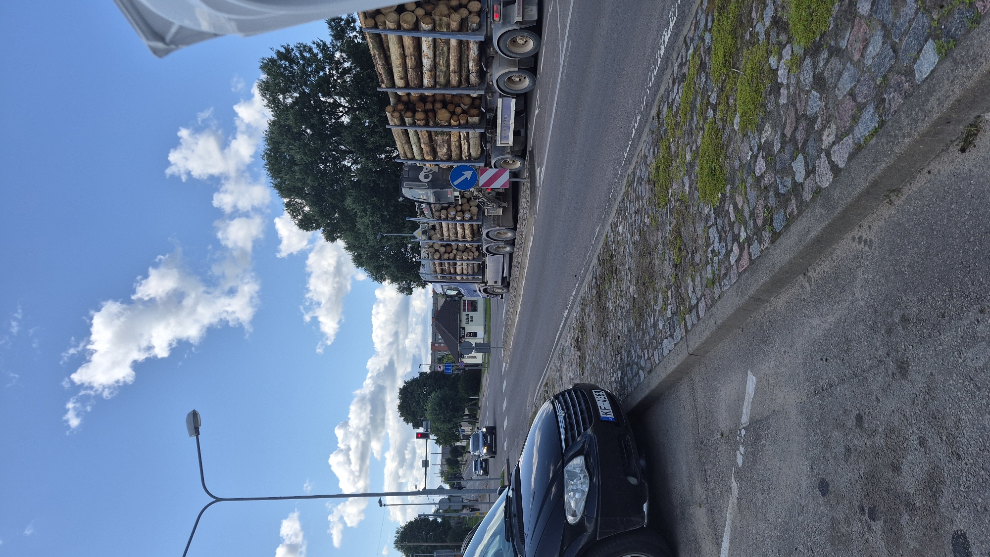 The image shows a sunny day with scattered clouds in Krāslava, Latvia. A black minivan is parked on the left side of the road, facing away from the viewer. The minivan's license plate reads "KF-4389". A large truck carrying logs is driving on the right side of the road, heading towards the viewer. The truck has a blue cab and a long trailer filled with logs. On the right side of the road, there is a small building with a sign that says "Kebab Bar". The road is paved with asphalt, and there is a sidewalk on the right side of the road made of cobblestones. In the background, there are trees and buildings.