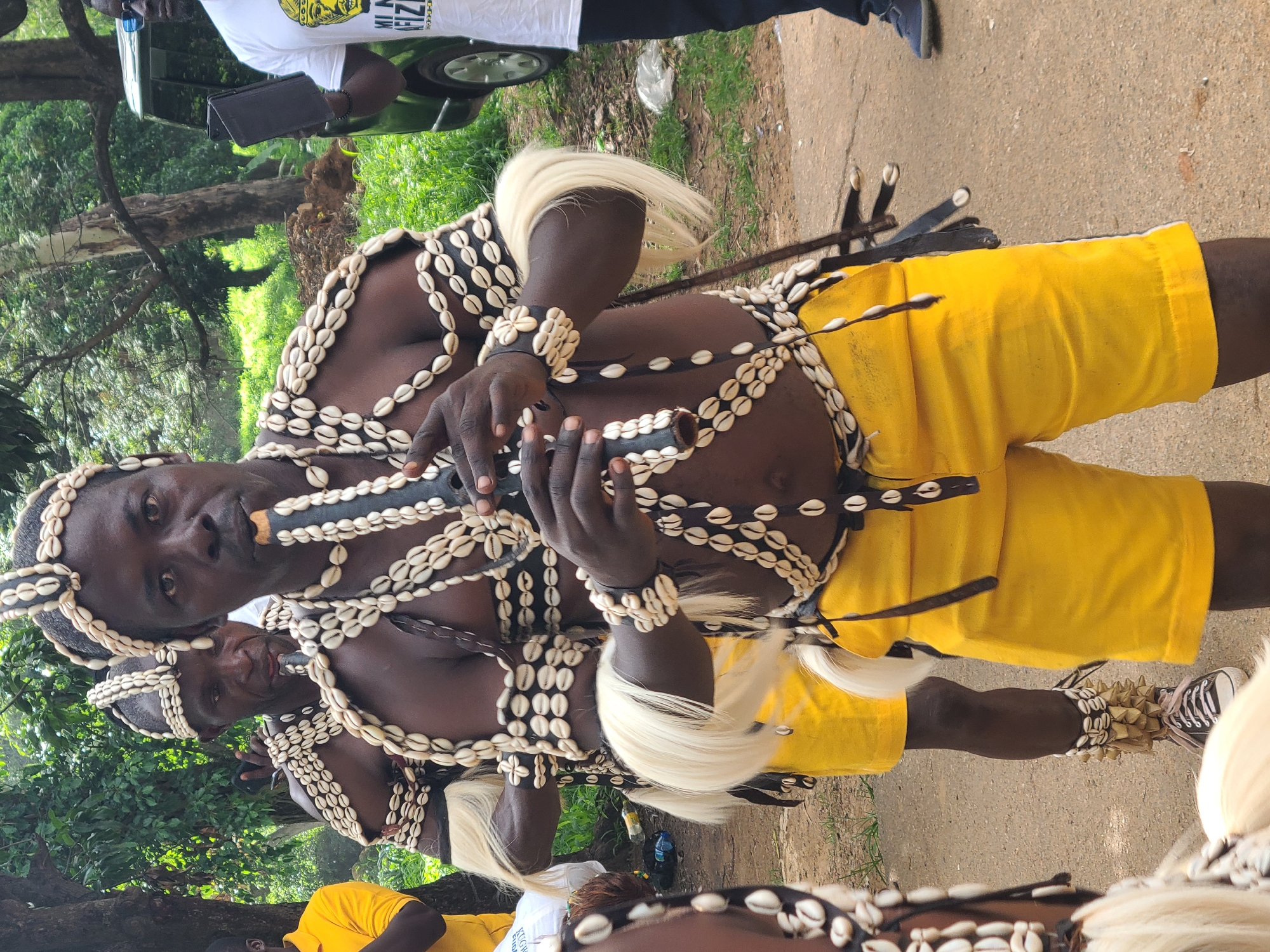 In Jos, Nigeria, three individuals are depicted in traditional attire. The foreground subject, a dark-skinned male with a stern expression, is playing a dark, cylindrical flute. He wears a headdress and chest adornments made of numerous white cowrie shells and black leather straps. His arms are adorned with cowrie shell bracelets and white faux fur accents. He is also wearing bright yellow shorts. To his left, another individual in similar traditional attire is partially visible, also holding a flute to his mouth. Behind them, a third person in a yellow polo shirt is partially visible on the far left. In the background, a dark-colored SUV is parked near lush green foliage and trees, suggesting an outdoor setting. The ground is a mixture of dirt and gravel. A man in a white t-shirt with a graphic print is standing to the right of the SUV, holding a black tablet.