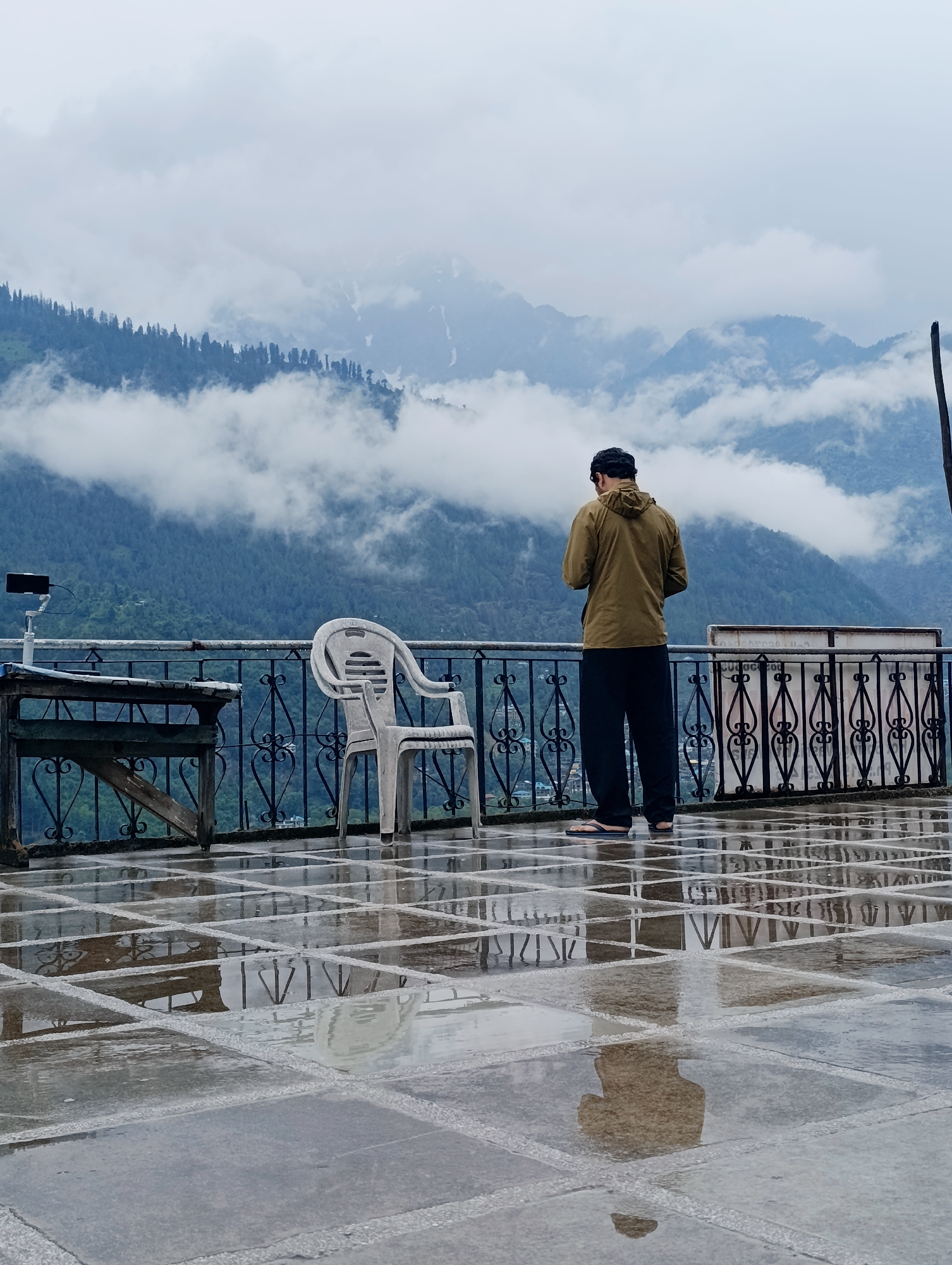 In Vashisht, India, on a cloudy day, a man stands on a wet, tiled patio overlooking a mountain range. The man, facing away from the camera, wears a light brown hooded jacket, dark pants, and flip-flops. He is holding a phone in his hands, looking down at it. To his left, a white plastic chair sits empty. A railing with decorative metalwork separates the patio from the forested mountains shrouded in mist and clouds. A table with a phone on a stand is also visible on the patio. The wet tiles reflect the railing, the chair, and the surrounding environment, creating a glossy surface.