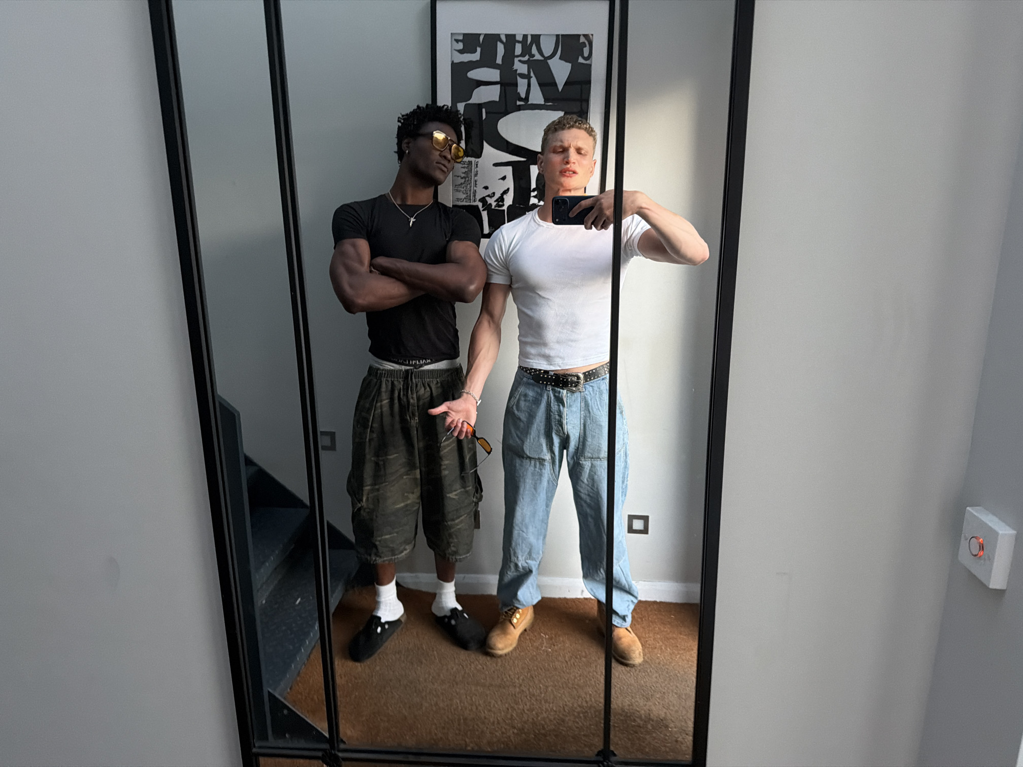Two men are reflected in a multi-paneled mirror. One man is Black, wearing dark-tinted sunglasses, a black t-shirt, camouflage shorts, and white socks with black shoes. He has his arms crossed. The second man is White, wearing a white t-shirt, light blue jeans with a black studded belt, and brown shoes. He is holding a smartphone and taking a photo of himself and the other man in the mirror.
In the background, there is a black and white art print hanging on the wall. To the left of the mirror, there is a staircase. To the right of the mirror is a white wall with a light switch. The location cannot be confirmed as being in London based on the image.