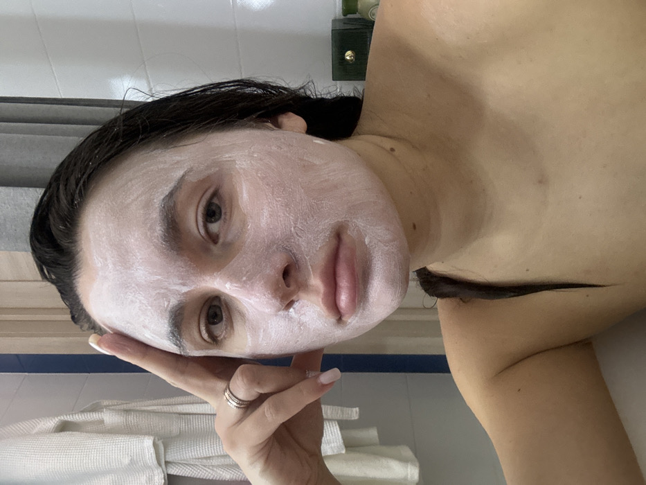 A fair-skinned woman lies on her side with a white face mask applied. She has dark hair and light brown eyes. Her left hand is raised near her face, revealing a silver ring. A stack of white towels is visible in the background, along with a blue and white tiled wall. Above her shoulder is a green and gold container.