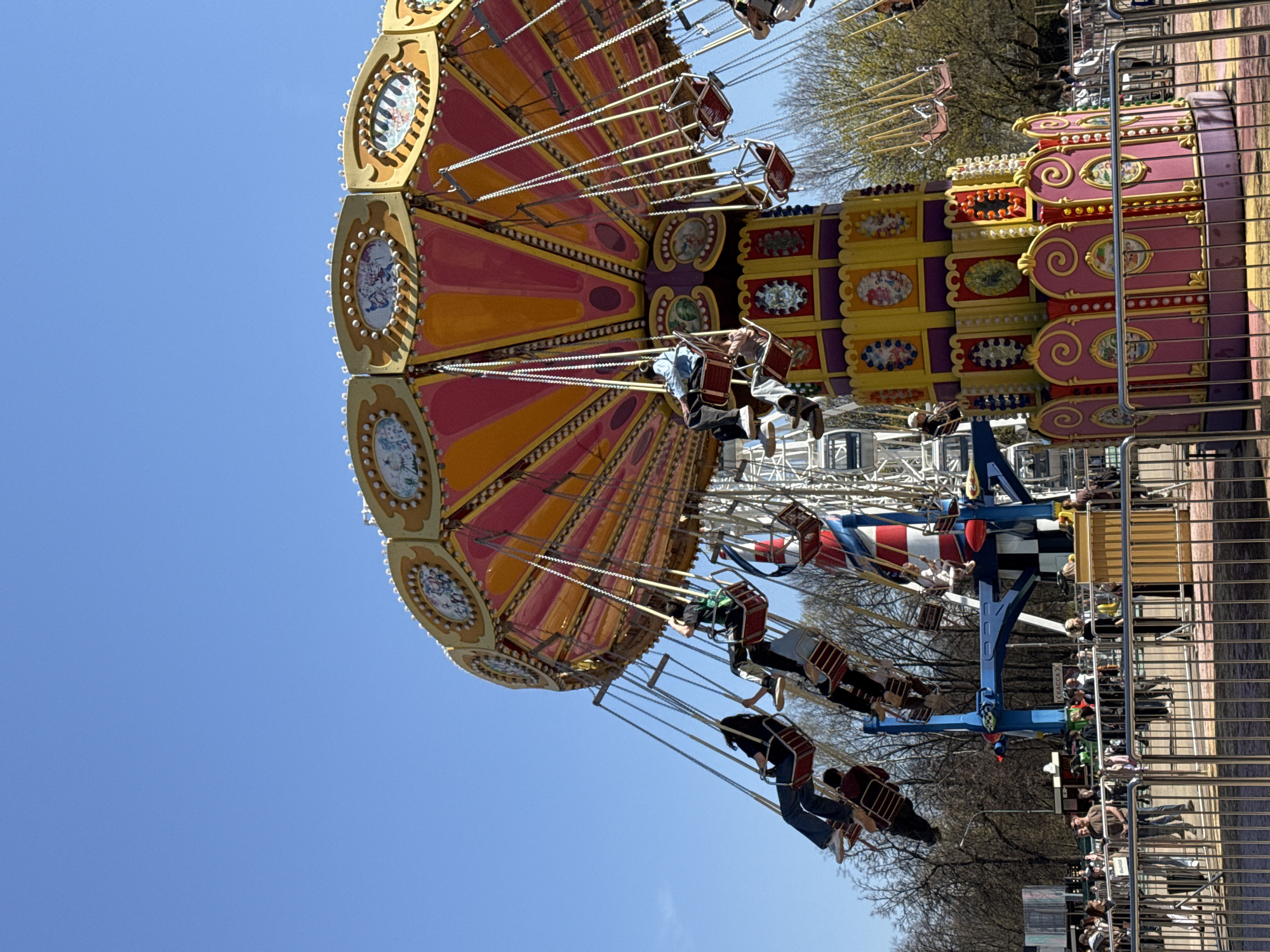 The image depicts a chair swing ride in motion, likely at a fair or amusement park. Riders are seated in suspended chairs swinging outwards due to centrifugal force. The ride structure is colorful, featuring hues of yellow, orange, pink, and purple, adorned with decorative patterns. The sky is clear and blue. People are visible on and around the ride, with some waiting behind a metal fence. The trees in the background lack leaves, suggesting it may be early spring or late autumn. Based on information available on the Internet, the amusement park ride appears to be located in Gorky Park in Moscow, Russia.
