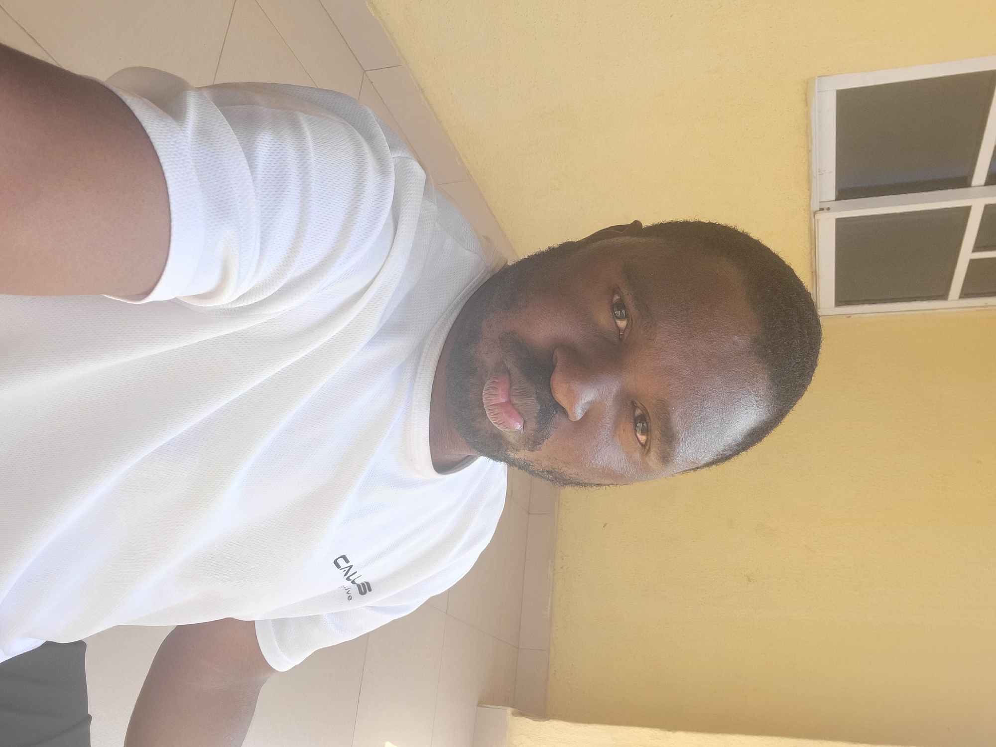 A medium shot, eye-level, selfie taken outdoors shows a Black man with dark skin, short black hair, and a beard puckering his lips as he looks at the camera. He is wearing a white, short-sleeved t-shirt with a small logo on the left chest. The man is positioned in the foreground, with his head and shoulders filling most of the frame. Behind him, the background consists of a light yellow wall with a window on the upper left. The window has white framing and appears to be closed. The ground in front of the wall is tiled with beige squares. The scene is likely on a patio or balcony, given the tiled flooring and the visible corner of a structure on the right. The lighting suggests it is daytime, with sunlight casting shadows. The location is identified as Suleja, Nigeria.