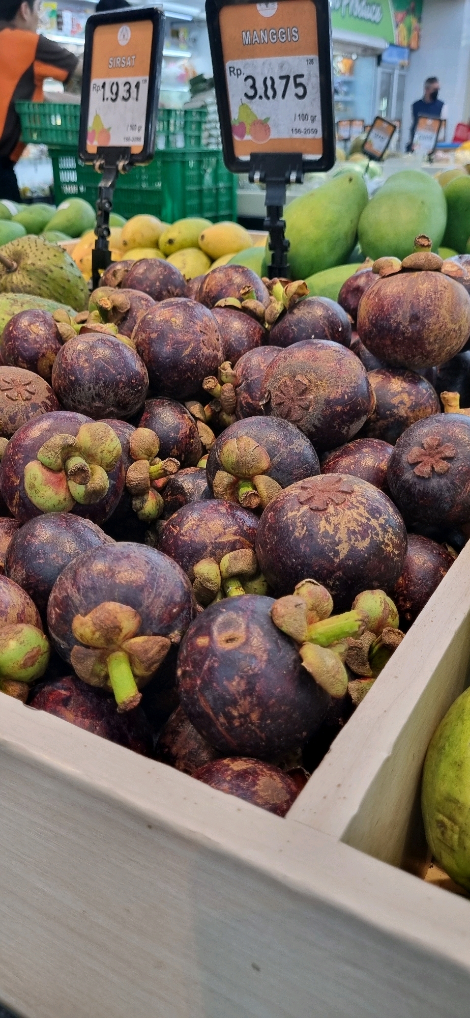 This image shows a close-up view of mangosteens and sirsak displayed at a market stall, likely in Denpasar, Indonesia. Two price tags are visible, one for "SIRSAT" priced at Rp. 1.931 per 100g, and another for "MANGGIS" priced at Rp. 3.875 per 100g. The mangosteens are dark purple and round, with prominent green calyxes. The sirsak, partially visible in the background, is green and spiky. In the background, other fruits such as mangoes are also present. Two individuals are partially visible; one is wearing an orange and black uniform, and another is wearing a dark shirt. The environment appears to be a well-lit indoor market.