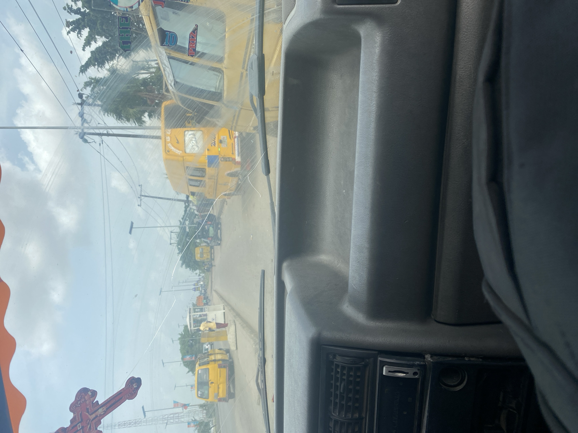 The image, rotated 90 degrees clockwise, captures a view from inside a vehicle through a heavily smudged and cracked windshield. The foreground shows the grey plastic dashboard on the bottom, with a windshield wiper blade resting horizontally across the center of the frame. On the bottom right, part of a vehicle console is visible, featuring a black vent and what appears to be a radio slot with "LOUDNESS" faintly etched.
Outside the vehicle, a paved road stretches into the distance under an overcast but bright sky. Several distinctive yellow buses or vans, often associated with public transport in West Africa (e.g., "danfo" in Nigeria), are visible. One prominent yellow bus is directly in front, bearing text like "20" and part of "NEW PRIZE" on its front. Another bus is further down the road, and more vehicles are scattered along the street.
Numerous utility poles and overhead power lines crisscross the sky. Green trees and foliage are visible in the background and along the roadside. On the far left, a large, ornate red and orange structure, possibly a monument, sign, or religious symbol (resembling a cross), is partially captured. Small flags on poles line the street in the mid-ground. Reflected on the windshield, the word "THE" is clearly visible, possibly part of a sign or banner outside. The overall scene suggests an urban or semi-urban environment during the daytime, likely in traffic or at a stop.