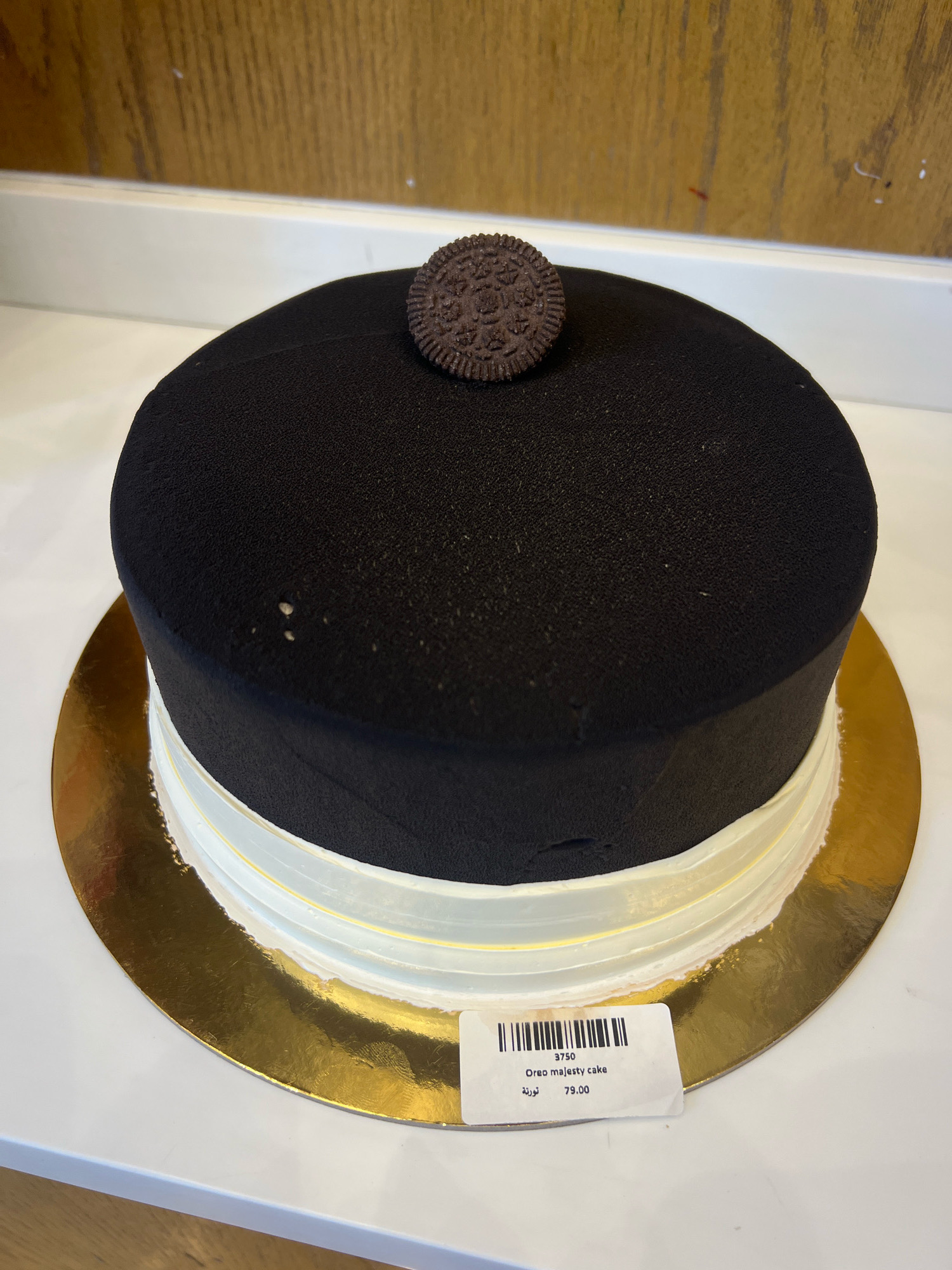 An Oreo Majesty cake is presented on a gold cake board, which rests on a white surface. The cake is round with a black, matte-textured frosting covering its top and sides. A single Oreo cookie is placed in the center of the cake's top. A white frosting band, edged with a yellow detail, encircles the base of the cake. A white price tag is affixed to the gold cake board, displaying "3750", "Oreo majesty cake", and "79.00" in English and Arabic script. The background shows a wooden textured wall and a white shelf. This cake is likely displayed in a bakery or patisserie in Al Khobar, Saudi Arabia.