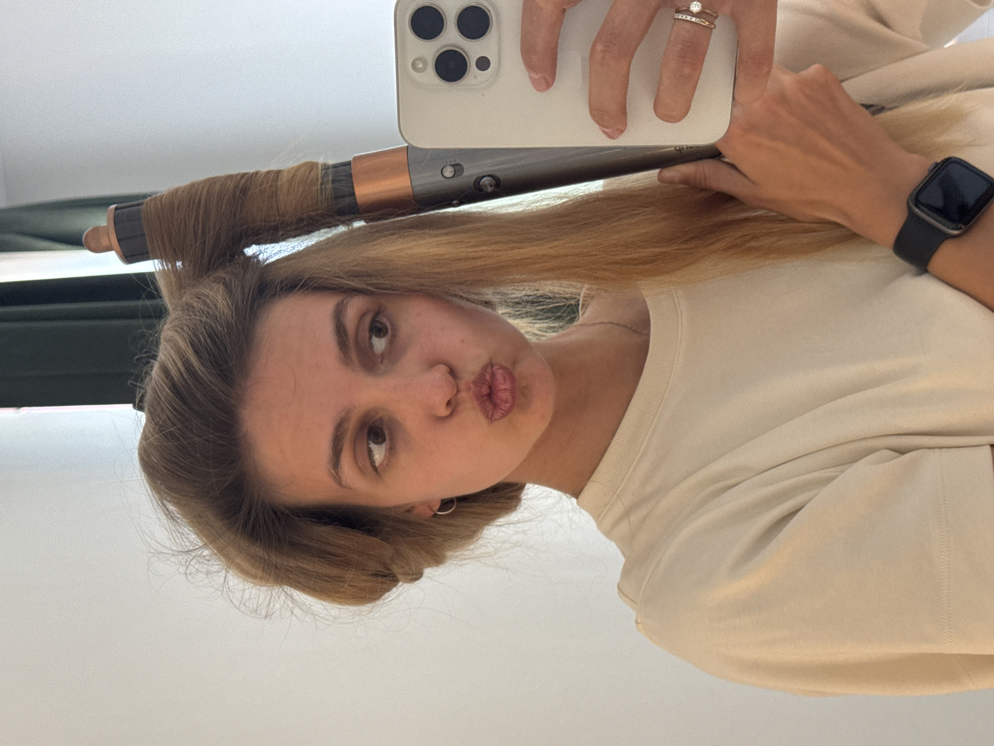 A woman is holding a hair curling iron to her hair in an indoor setting. She is wearing a light-colored shirt and an Apple Watch on her left wrist. She is also holding a white iPhone in her right hand, presumably taking the picture. The location is Unknown, Unknown.
