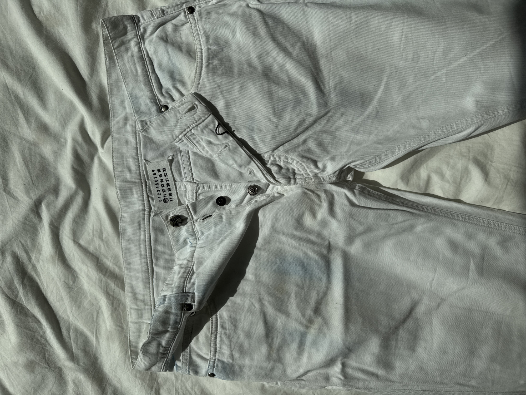 A pair of light-colored denim trousers is displayed in a flat lay, likely indoors in London, United Kingdom. The jeans, possibly off-white or very light blue, are laid out on a wrinkled, lighter-toned fabric surface, possibly a bedsheet. Strong natural light, presumably sunlight from a window, illuminates the scene from the upper left, casting prominent shadows that highlight the folds and textures of the fabric.
The trousers feature an open button-fly closure with multiple dark-colored metal buttons. The fabric appears slightly worn or distressed, with some subtle bluish discoloration on the upper front thigh area, consistent with denim wear or a specific wash.
A rectangular white fabric tag is sewn onto the left side of the waistband, displaying numbers arranged in rows. The visible text reads:
"0 1 2 3 4 5 6 7 8 9"
"10 11 12 13 14 15 16"
"17 18 19 20 21 22 23"
The number '11' is distinctly circled on this tag. The presence of strong shadows suggests it is either late morning or afternoon on a clear day. No specific activity is taking place; it is a static product shot.