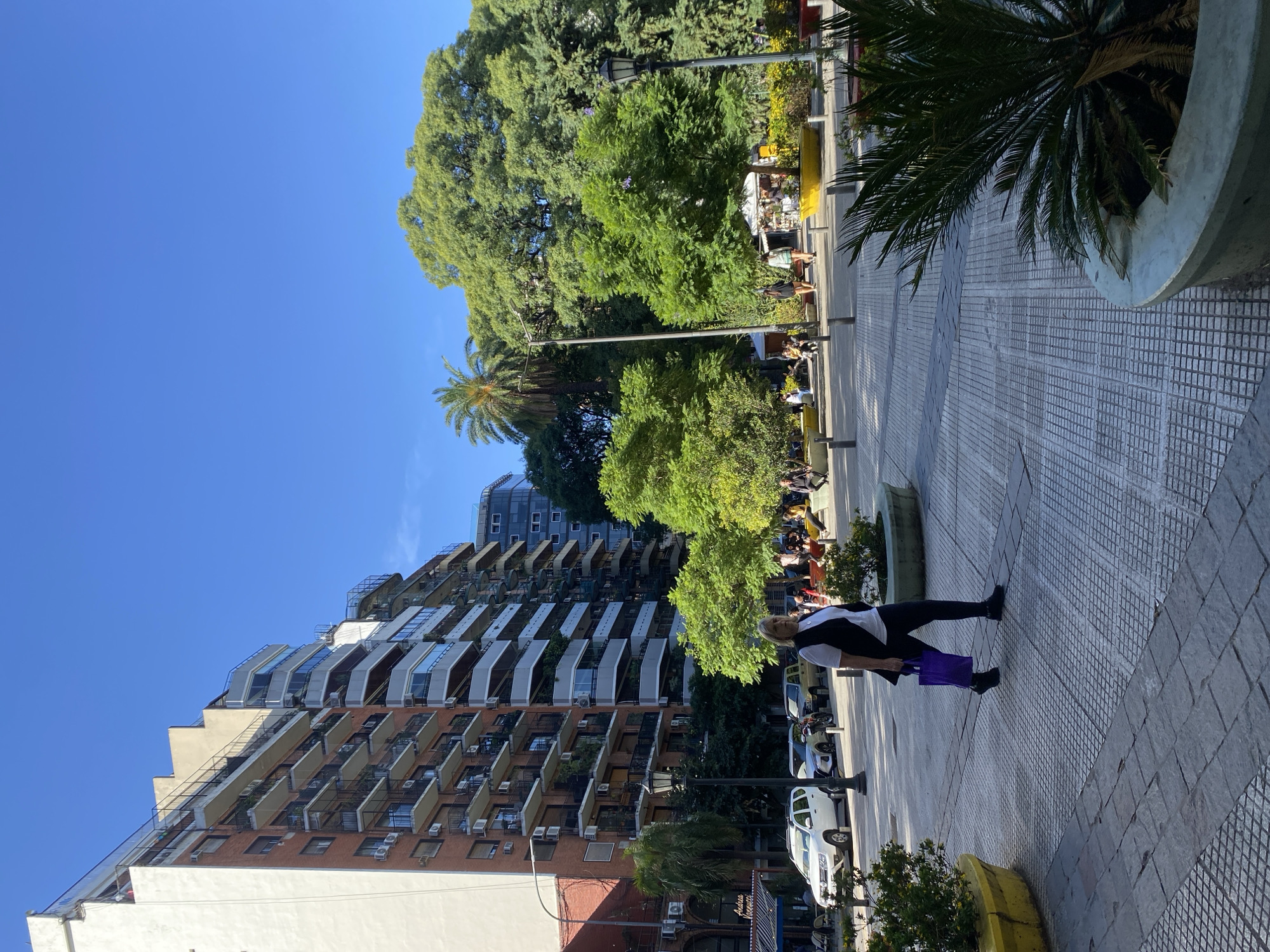 The image captures a scene in Buenos Aires, Argentina. A tall, modern apartment building with numerous balconies dominates the left side of the image. A woman is walking along a paved sidewalk towards the viewer. She wears a white shirt, a dark vest, black leggings, and carries a purple bag. Behind her is a row of parked cars, a park with trees and people sitting on benches. The sky is clear and blue.