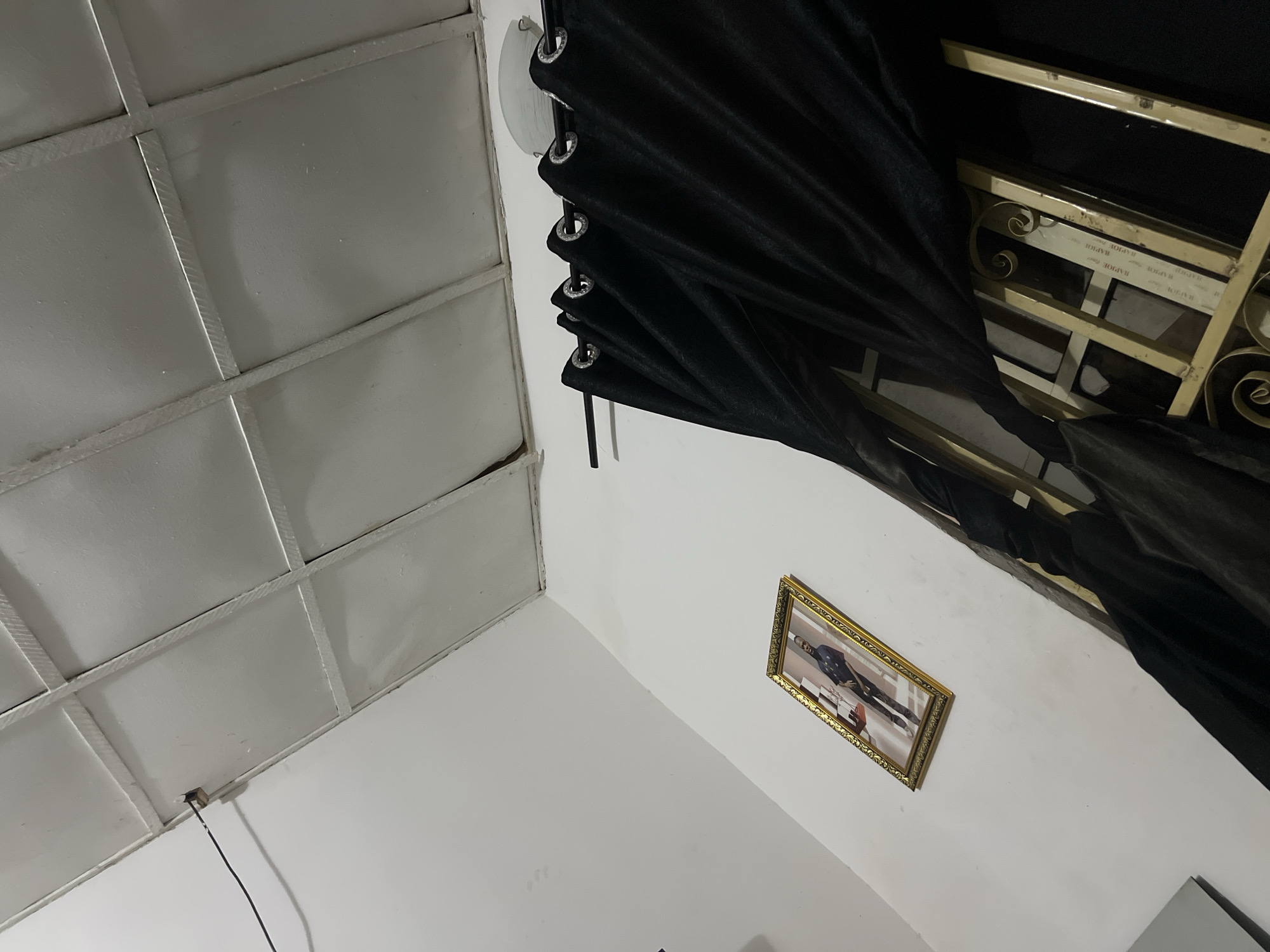 The image depicts an indoor scene, possibly a room in Howayi, Nigeria. A framed portrait of a man in uniform hangs on a white wall. To the right, black curtains with grommets are drawn across a window, partially obscuring ornate metal bars. The ceiling, made of white panels, shows a grid pattern. A black electrical cord is visible near the ceiling, attached to the wall with a small fixture. The lighting appears dim.