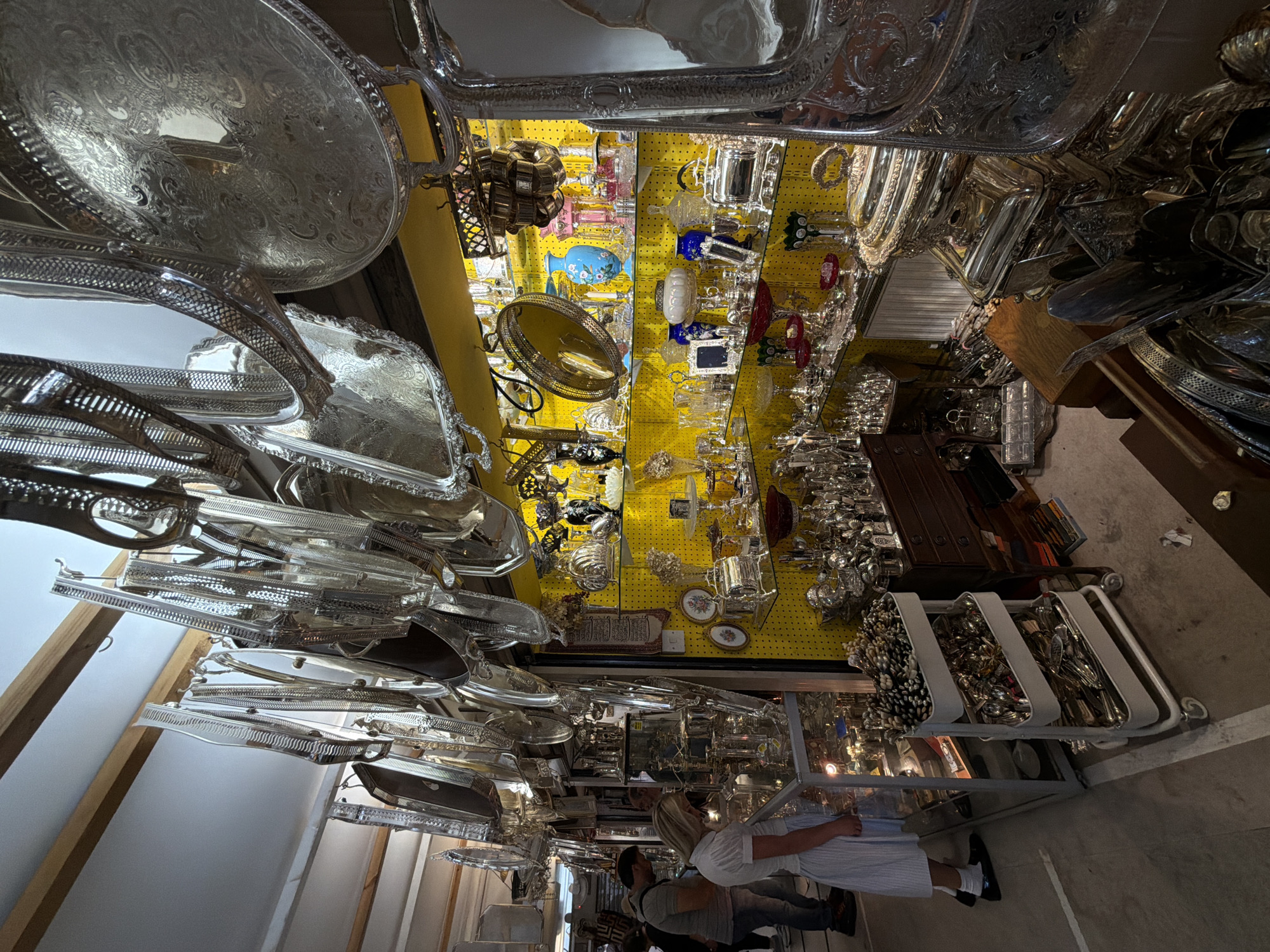 The image depicts the interior of an antiques shop, potentially located in London, United Kingdom, given the abundance of vintage and ornate silver items. The space is densely packed with merchandise displayed on shelves and racks, including silver platters, serving dishes, glassware, and decorative objects. The shop has a distinctive yellow pegboard wall, upon which a variety of items are hanging, and a display case containing silverware. Several customers are present, browsing the items. The lighting is a combination of natural light and artificial illumination, creating a somewhat cluttered and atmospheric environment.