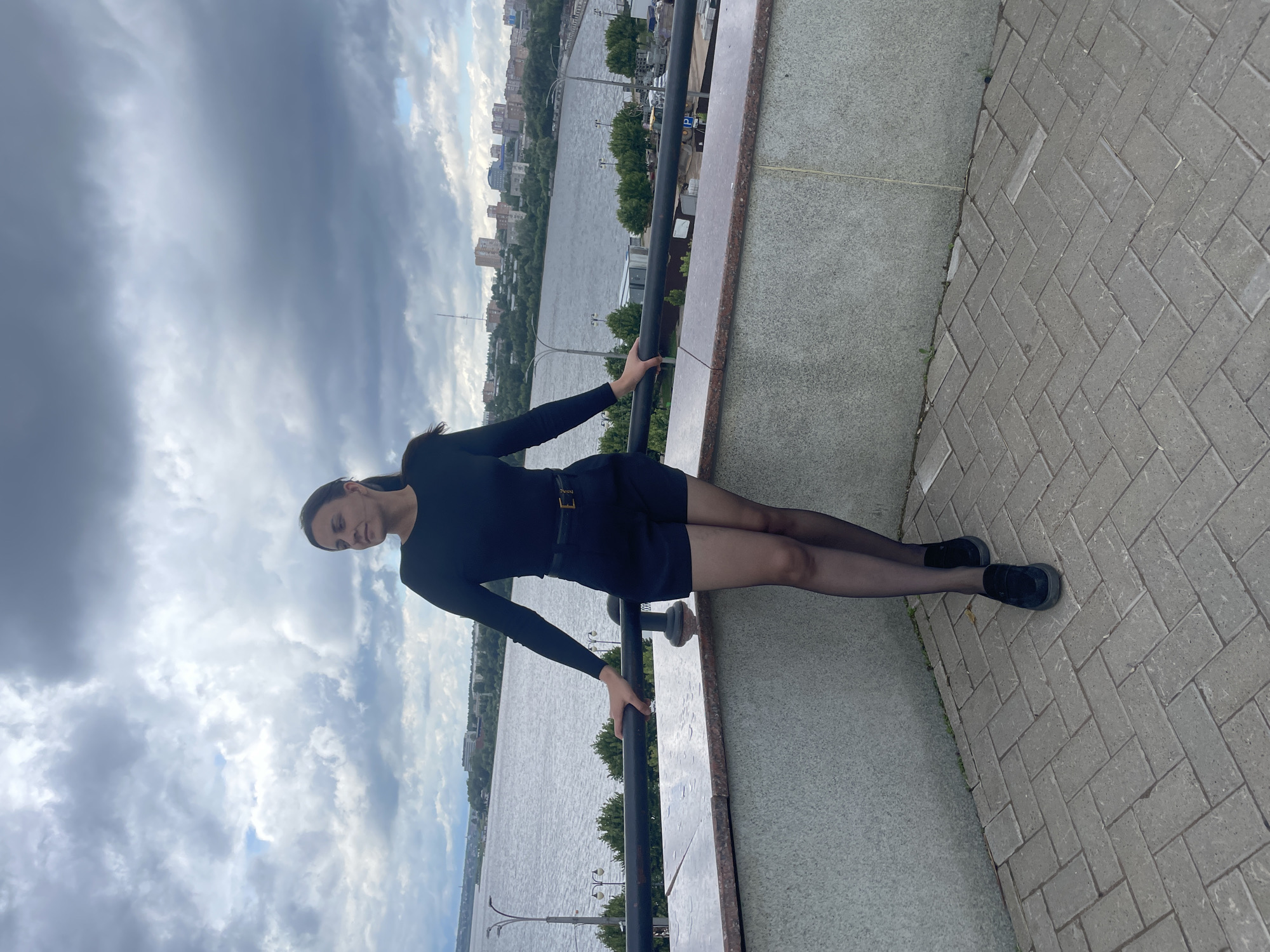 A full-body, slightly elevated shot captures a woman standing on a paved balcony overlooking a city and a body of water under a cloudy sky. The woman, facing left and looking away from the camera, wears a black long-sleeved top, black shorts with a belt, black sheer tights, and black loafers. Her left hand rests on a dark railing, while her right arm is bent at the elbow. The balcony features a patterned brick floor and a granite-patterned border. In the background, a wide river separates the balcony from the city of Izhevsk, Russia, with buildings lining the opposite bank. The sky is filled with dramatic, dark clouds, suggesting overcast weather.