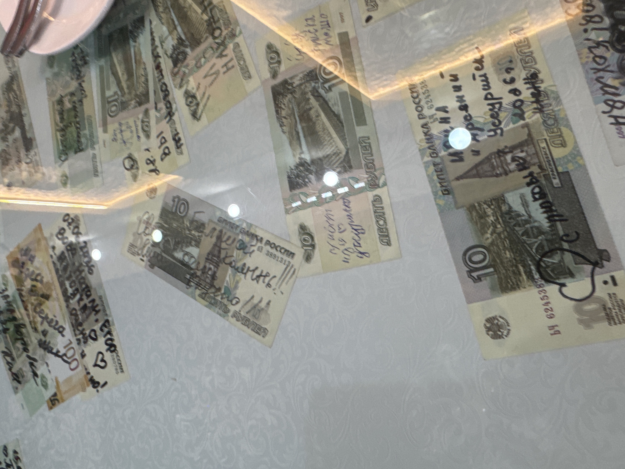 The image shows several Russian banknotes scattered on a white, patterned surface, possibly a tablecloth. The banknotes are marked with handwritten messages and drawings in black ink. The messages appear to be in Russian and include names, dates, and personal messages. Some banknotes also have drawings, such as hearts. The lighting suggests an indoor setting, with reflections from overhead lights visible on the surface. There are no people visible in the image. The banknotes are of various denominations, including 10 and 100 rubles. The surface has a subtle floral or damask pattern. The banknotes are oriented at various angles, and some are partially obscured by others. The overall impression is of a casual or informal setting where these banknotes have been left or used. There is no discernible information within the image that would indicate the location in Suifenhe, China.