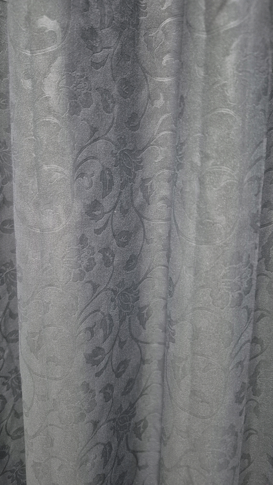 A grey, patterned curtain hangs in a room. The curtain features a damask-like floral pattern in a slightly darker shade of grey. The fabric is gathered, creating vertical folds and shadows. The lighting appears to be ambient, casting soft shadows on the textured surface. The location in Gambiranom, Indonesia is not discernable from the image.