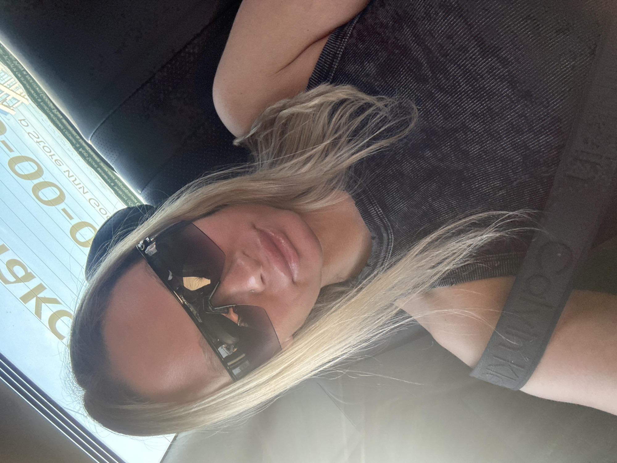 A blonde woman wearing sunglasses and a black t-shirt with a Calvin Klein strap is photographed in Perm, Russia. The reflection in the sunglasses shows the Perm city landscape with buildings and lettering in reverse in Russian which reads “Nikolin company”. The scene appears to be inside a vehicle.