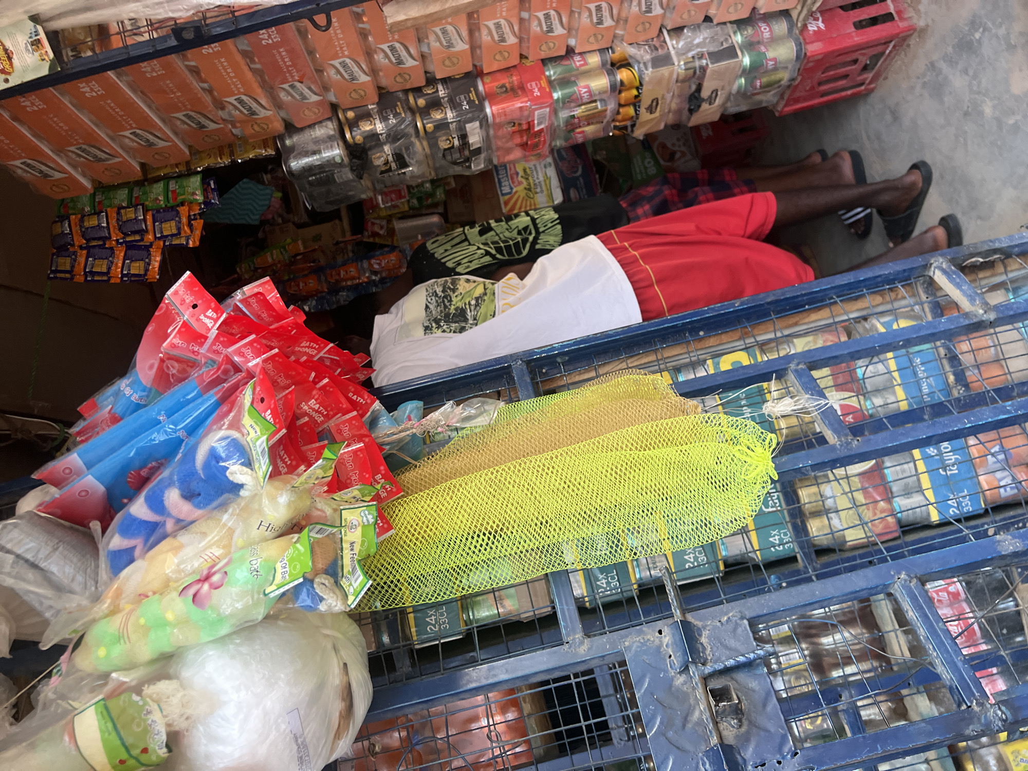 This image captures the densely packed interior of a convenience store or kiosk in Gbaza, Nigeria. Two individuals are partially visible from the back within the narrow shop space. On the left, a person wears a white t-shirt with a dark graphic and red shorts. Behind and to their right, another person wears a black t-shirt with a green graphic and plaid shorts, along with black slide sandals. A third pair of legs, similarly dressed in plaid shorts and black slide sandals, is visible further back on the right.
The shop is extensively merchandised. A prominent blue metal gate or grating secures the left side of the frame. Various bath sponges or loofahs in colorful plastic packaging (green, yellow, blue, red) are suspended from above and to the left, alongside a white plastic bag and a bright yellow mesh bag. The right side of the shop is dominated by vertical stacks of packaged beverages. These include multiple cases of "MALTINA NON-ALCOHOLIC MALT DRINK" (24x 33CL cartons) and stacks of "FEARLESS ENERGY DRINK" (black and gold cans, 24x 250ml). Other assorted beverage cans, including Fanta and Coca-Cola, are visible behind the blue grating. Small packets of snacks or candies in orange and green packaging hang from strings in the upper center of the shop. The floor is concrete, and red plastic crates are used for stacking merchandise on the lower right.
